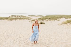 A person in a light blue dress walks along sandy beach dunes on a sunny summer day.