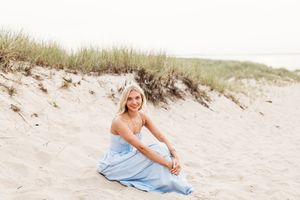 A person in a light blue dress poses gracefully on a sandy beach with coastal grass in the background.