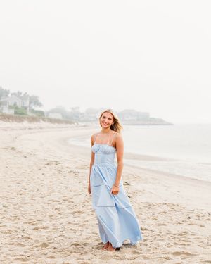 A woman in a light blue sundress walks along a sandy beach on a bright overcast day.