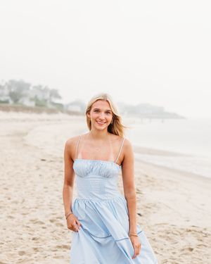 Woman in a light blue sundress poses on a sandy beach with ocean waves and cloudy sky in the background.