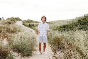 A person in white clothing walks through tall beige grass on rolling coastal sand dunes on a sunny day.