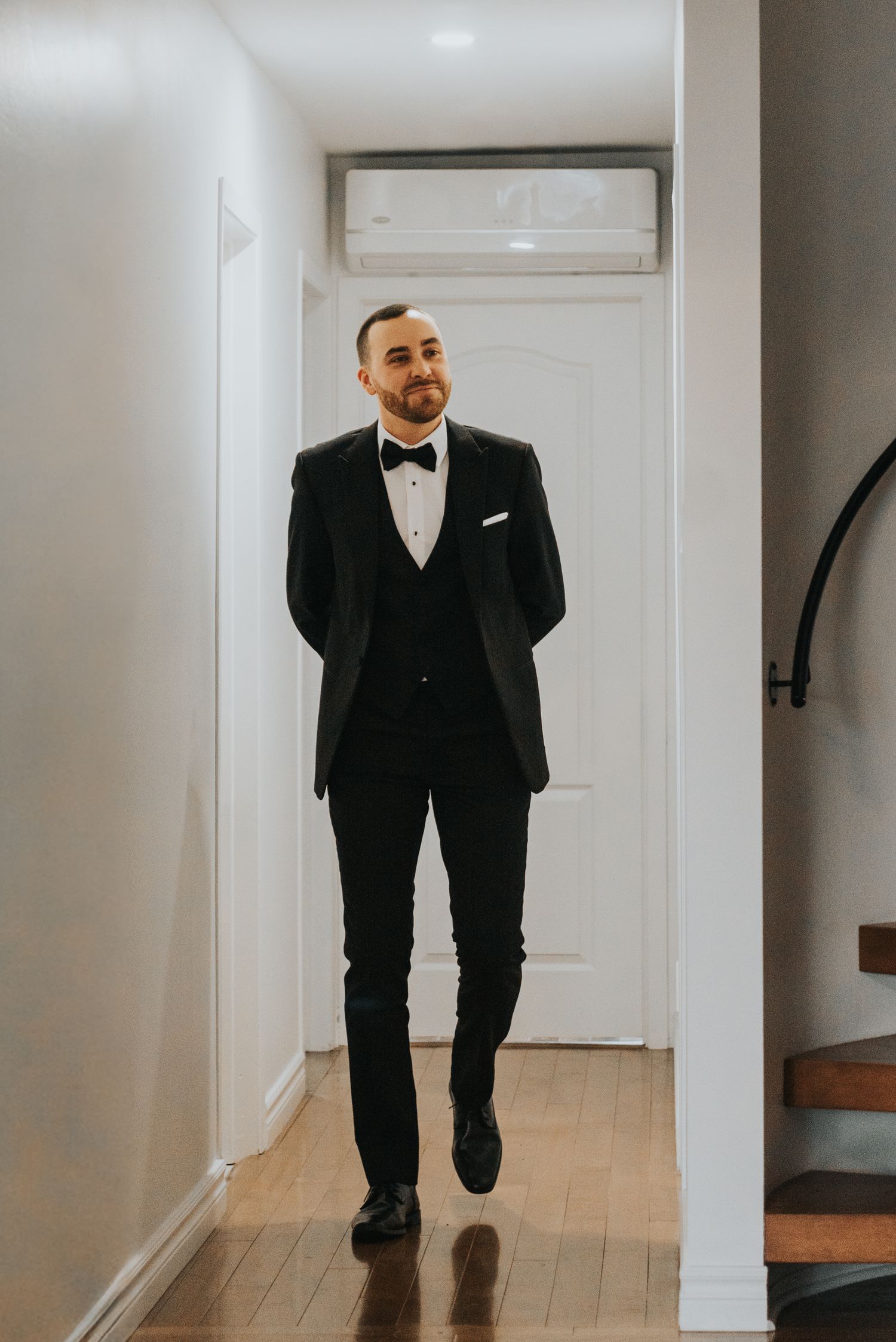A person in a classic black tuxedo and bow tie walks down a bright hallway with wooden floors.
