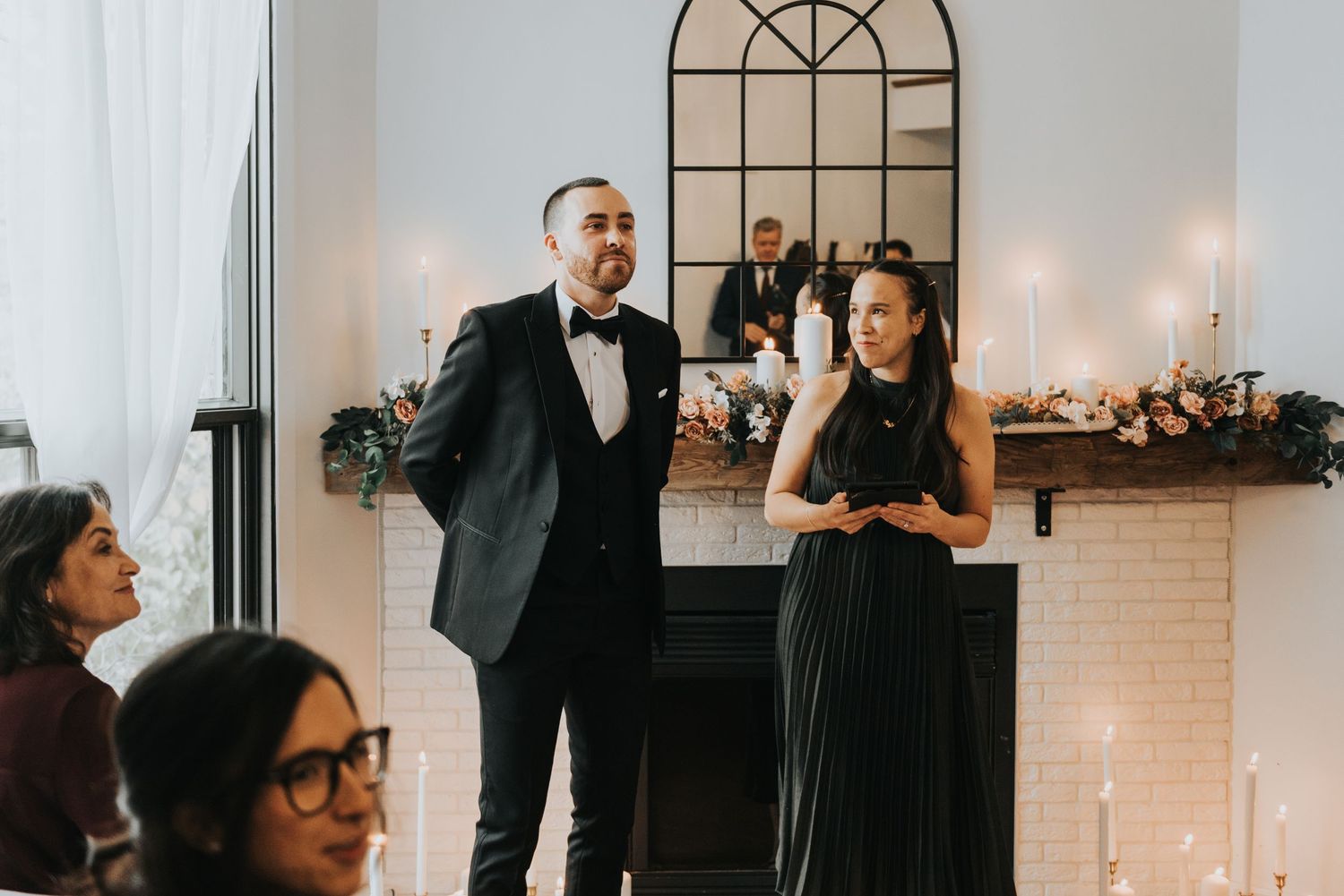 Wedding reception scene with guests gathered around a white brick fireplace decorated with candles.