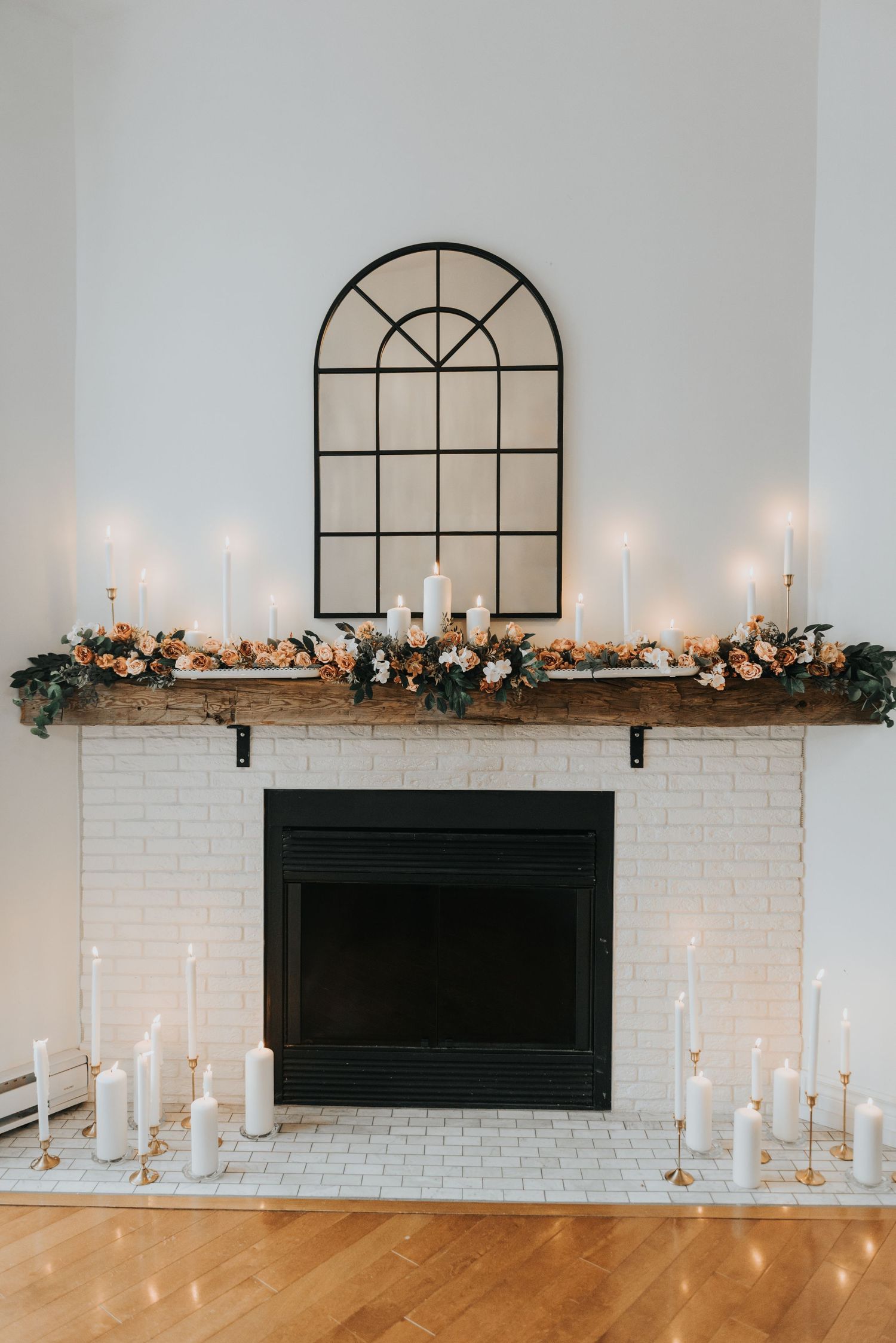 White brick fireplace decorated with elegant floral garland and white candles beneath an arched window frame.