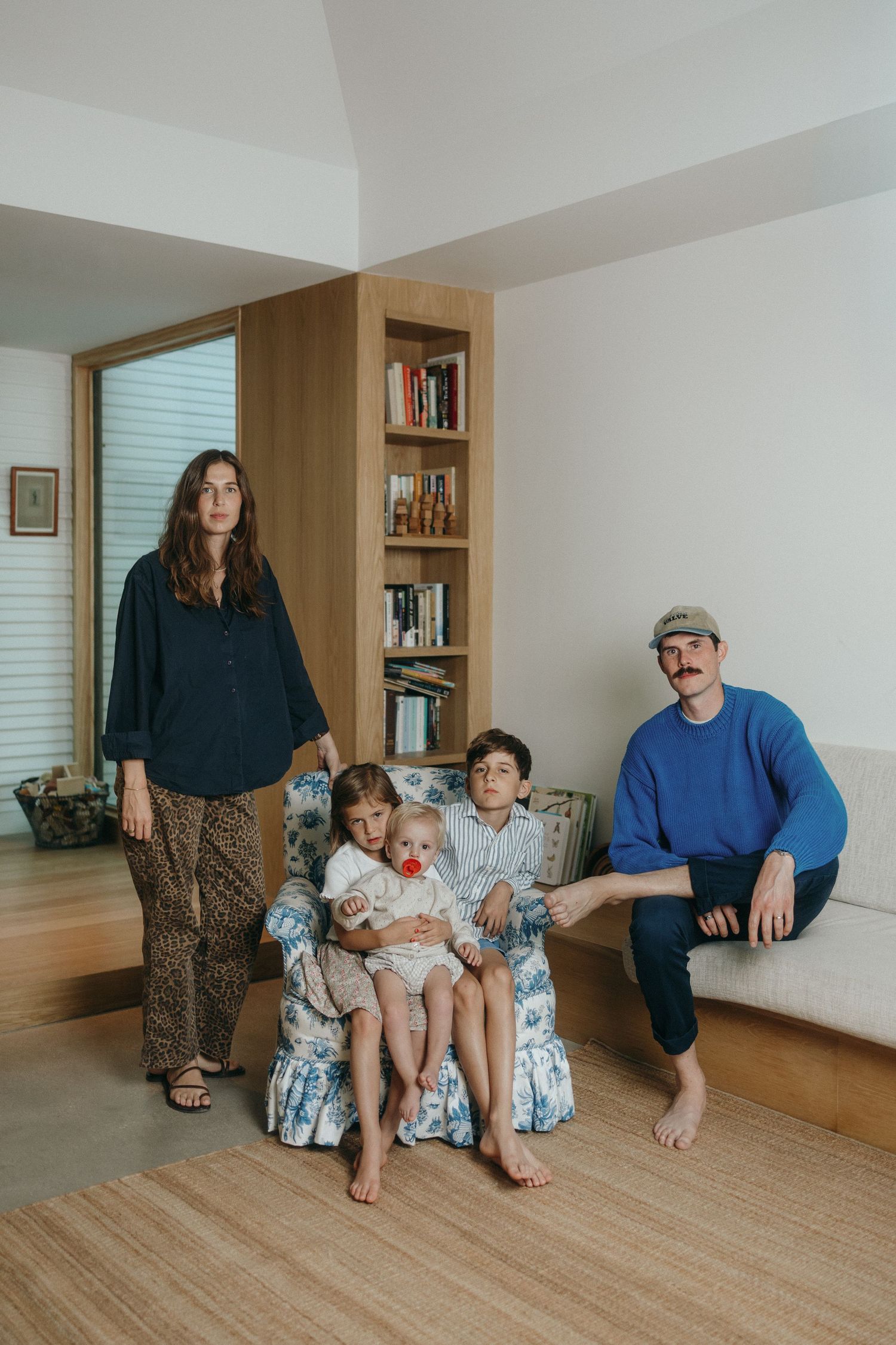 Family sits together on wooden floor in bright minimalist room with built-in bookshelves.
