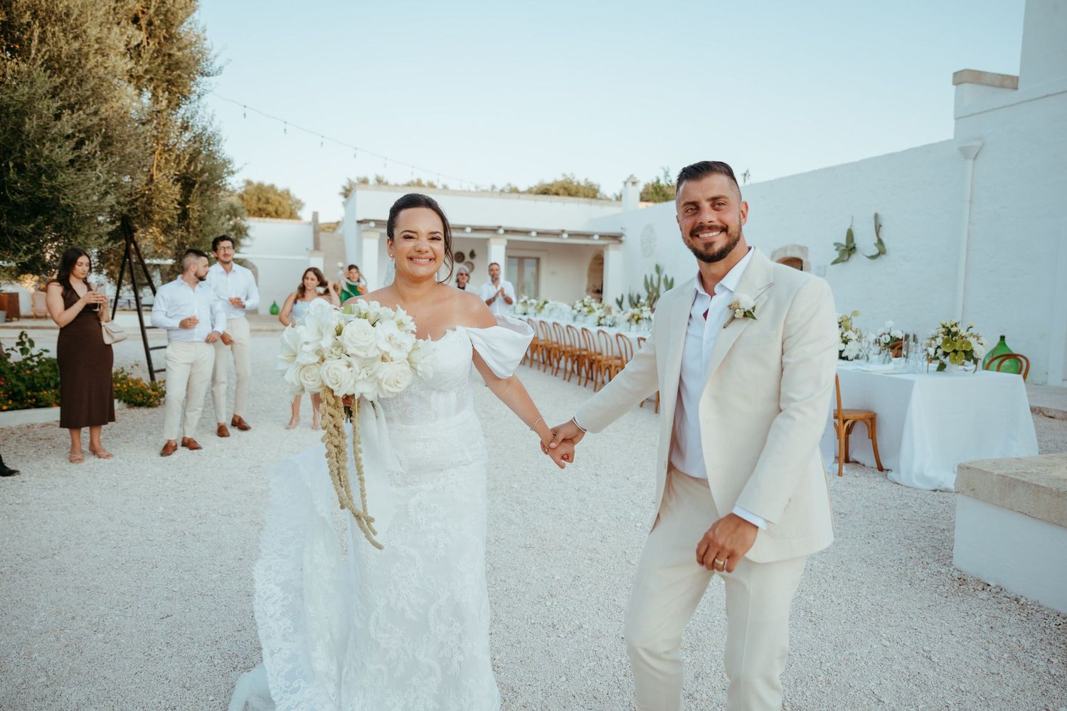 A couple celebrates their wedding day in white attire at an outdoor Mediterranean venue during sunset.