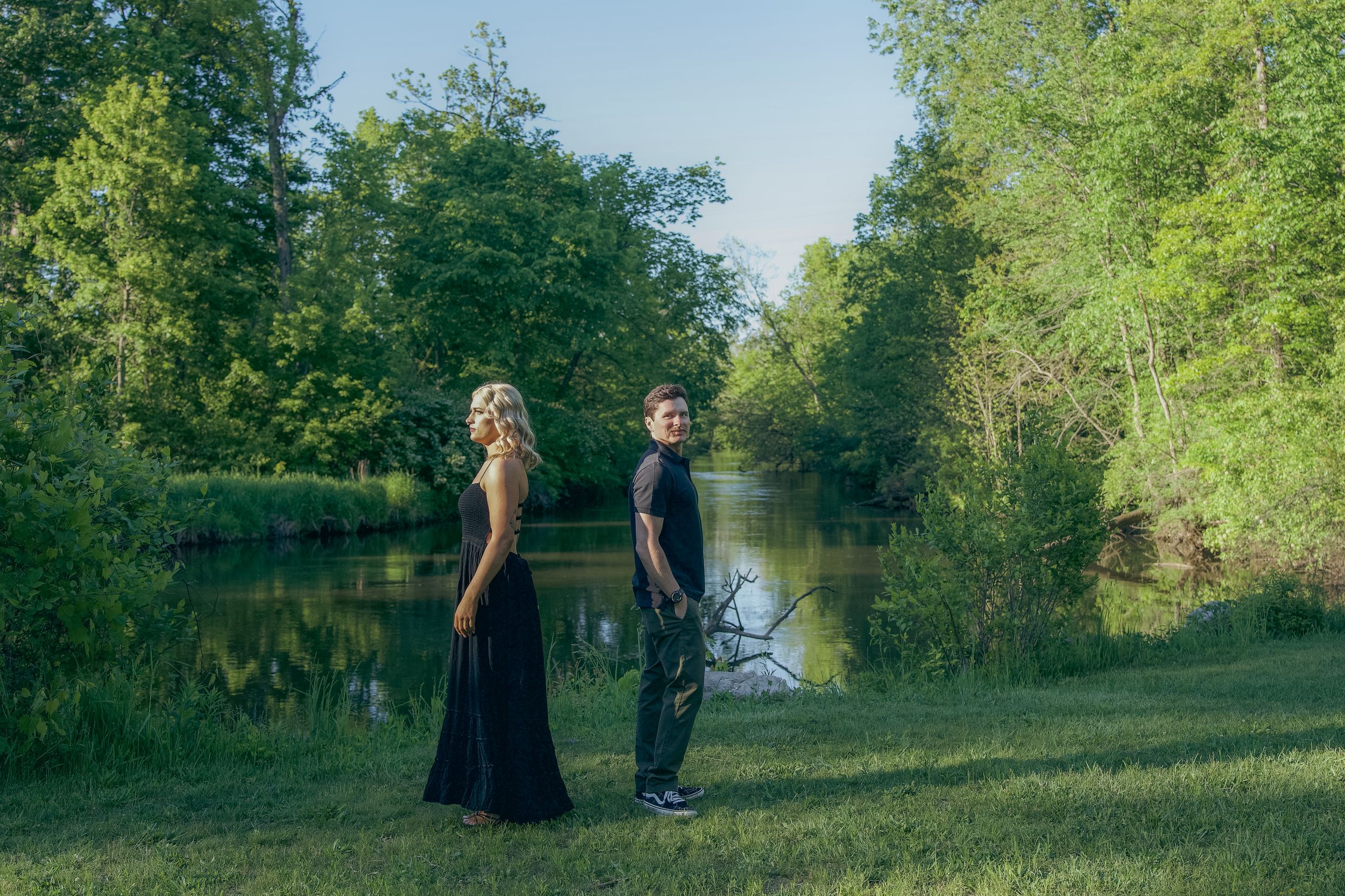 Two people stand on a grassy bank beside a tranquil pond surrounded by lush green trees on a sunny day.
