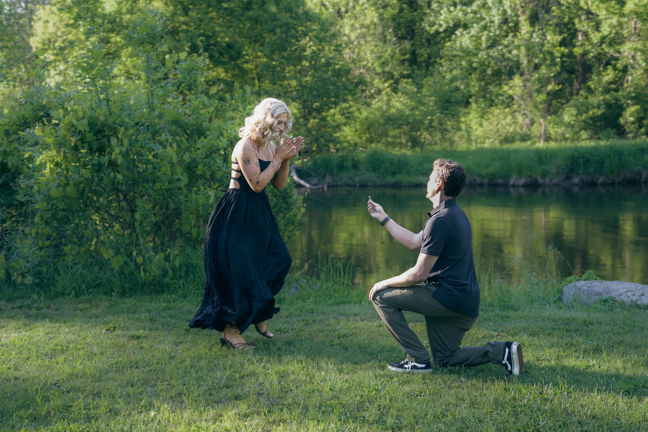 Two people sit together by a tranquil lake surrounded by lush greenery on a sunny afternoon.