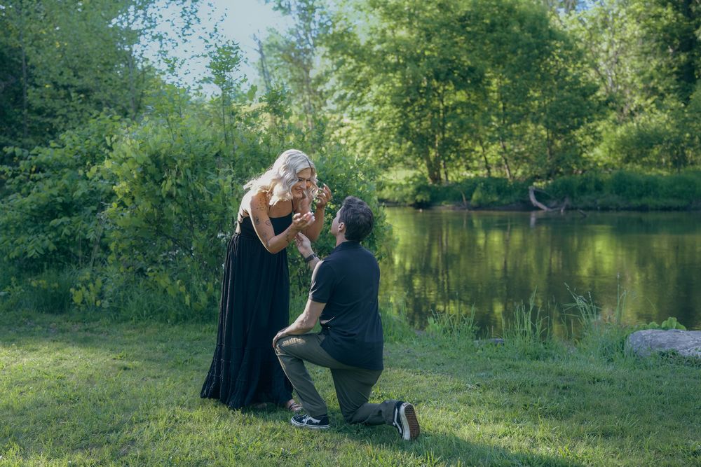 A romantic proposal scene unfolds by a serene pond surrounded by lush greenery on a sunny afternoon.