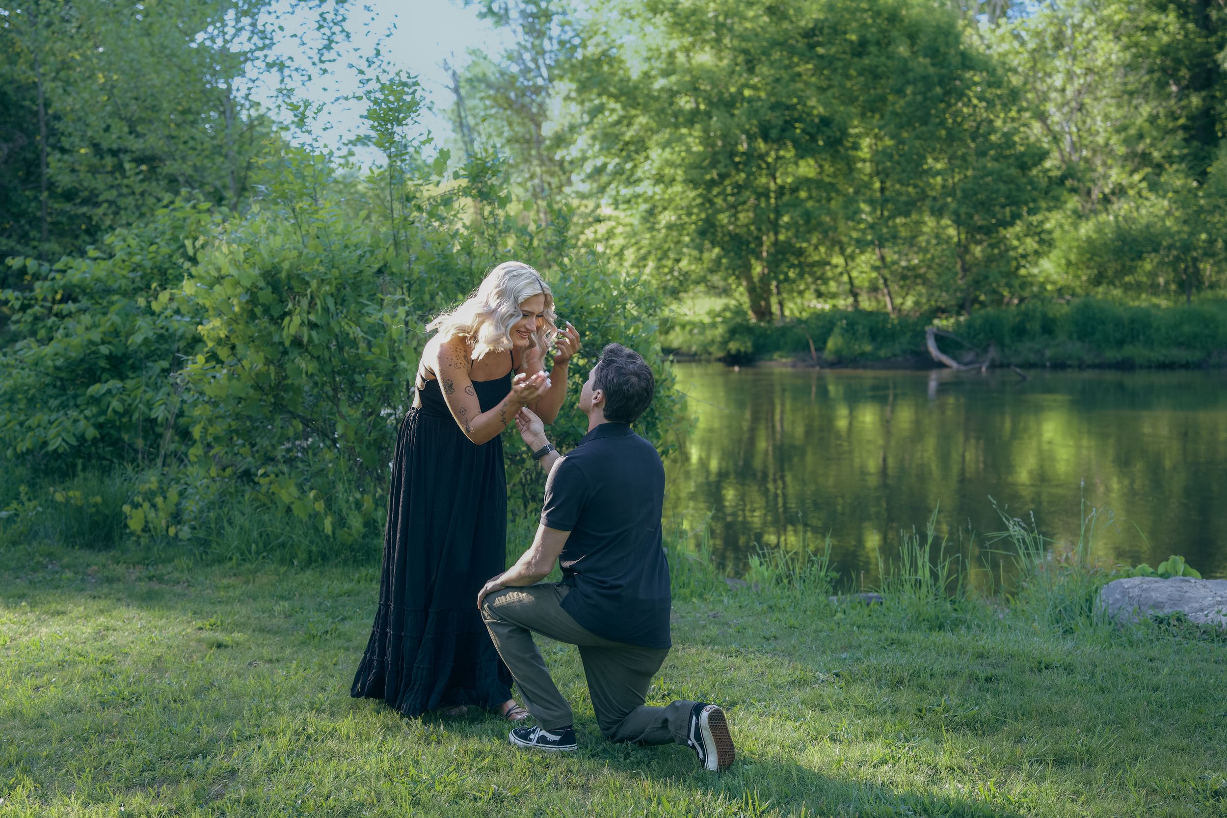 A romantic proposal scene unfolds by a serene pond surrounded by lush greenery on a sunny afternoon.