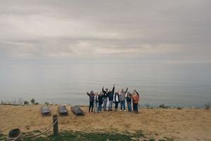 A group of friends raise their arms in celebration while standing on a cliff overlooking a serene ocean under cloudy skies.