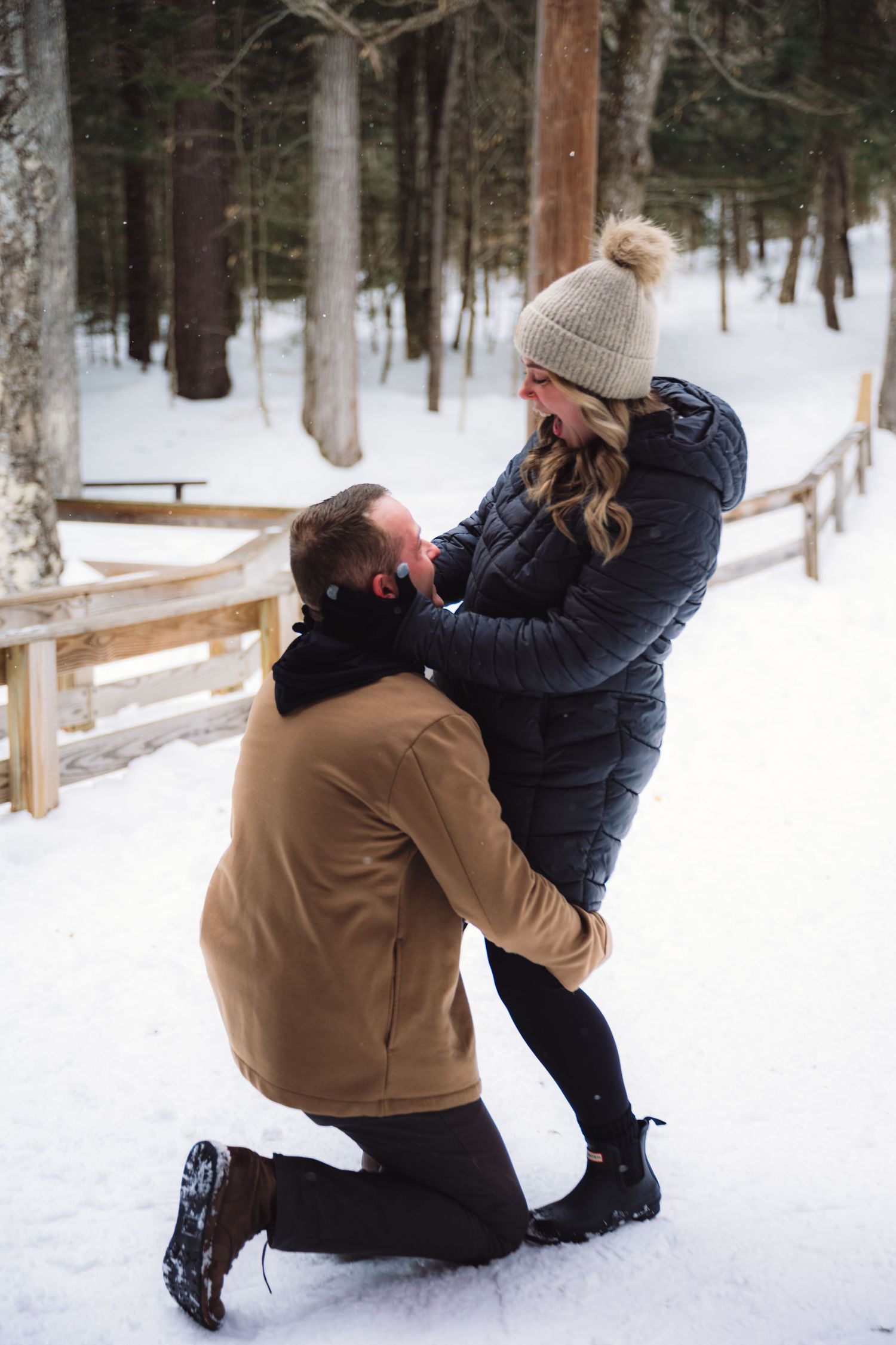 Surprise winter engagement photo in Lincoln, New Hampshire featuring a couple kneeling in fresh snow along a rustic wooden fence