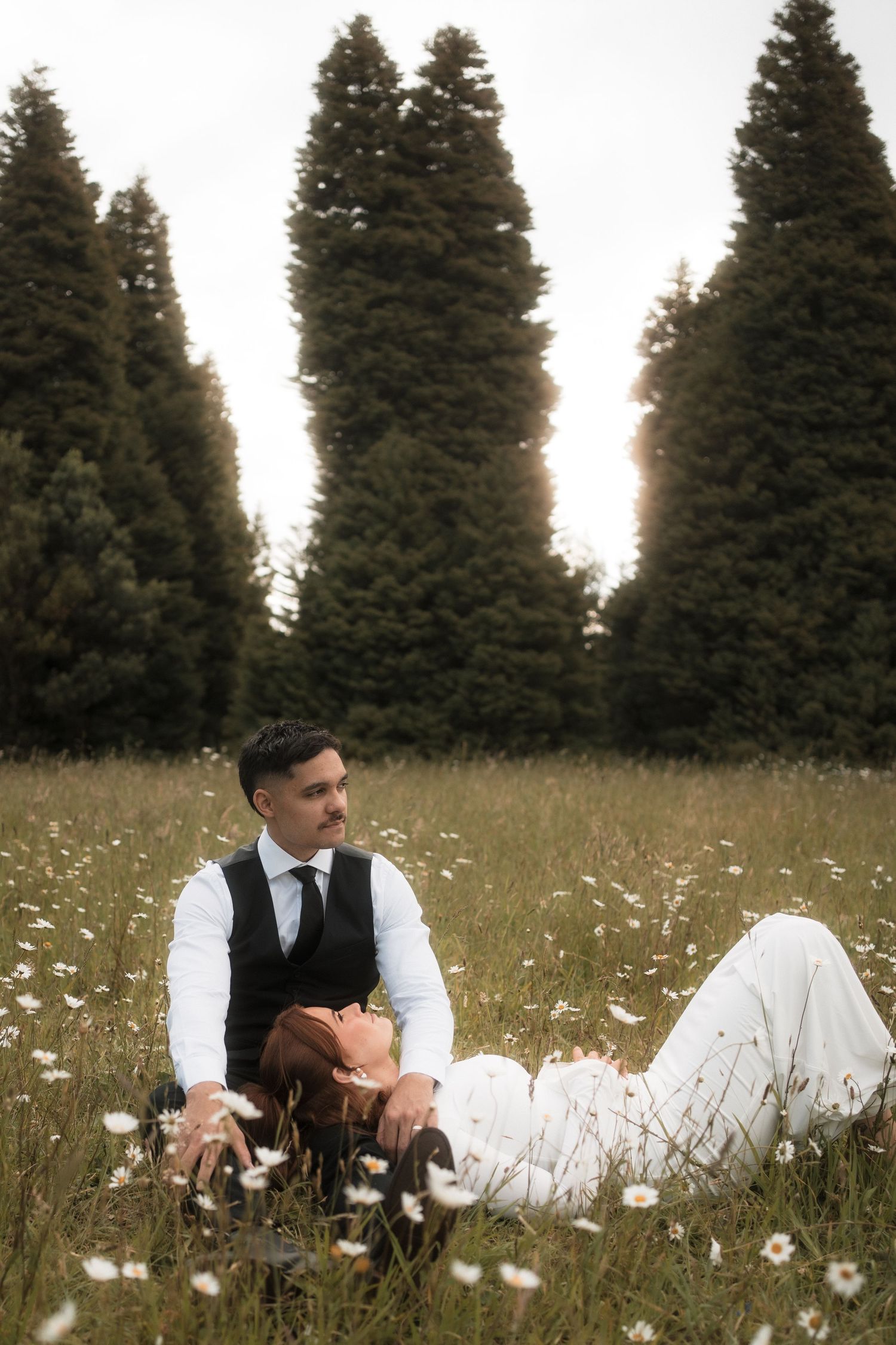 A couple in formal attire sits in a wildflower meadow surrounded by tall evergreen trees at dusk at RJ Hamer, Olinda