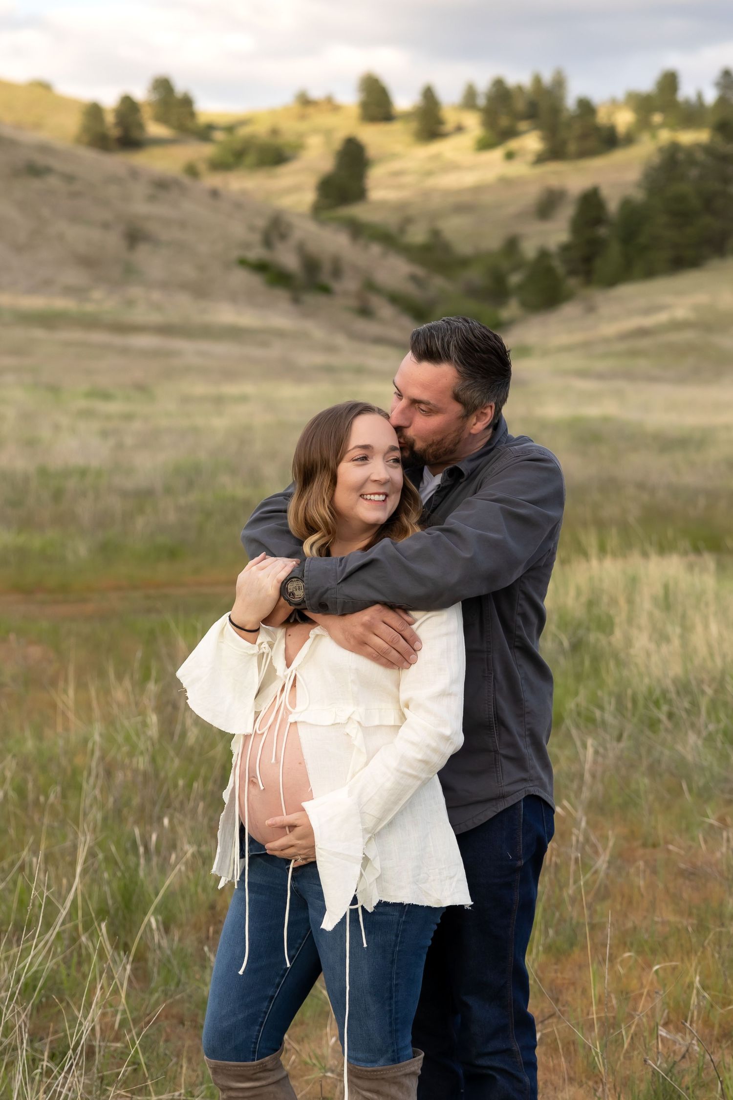 A couple embraces playfully in a scenic field with rolling hills and tall grass during a romantic outdoor photoshoot.