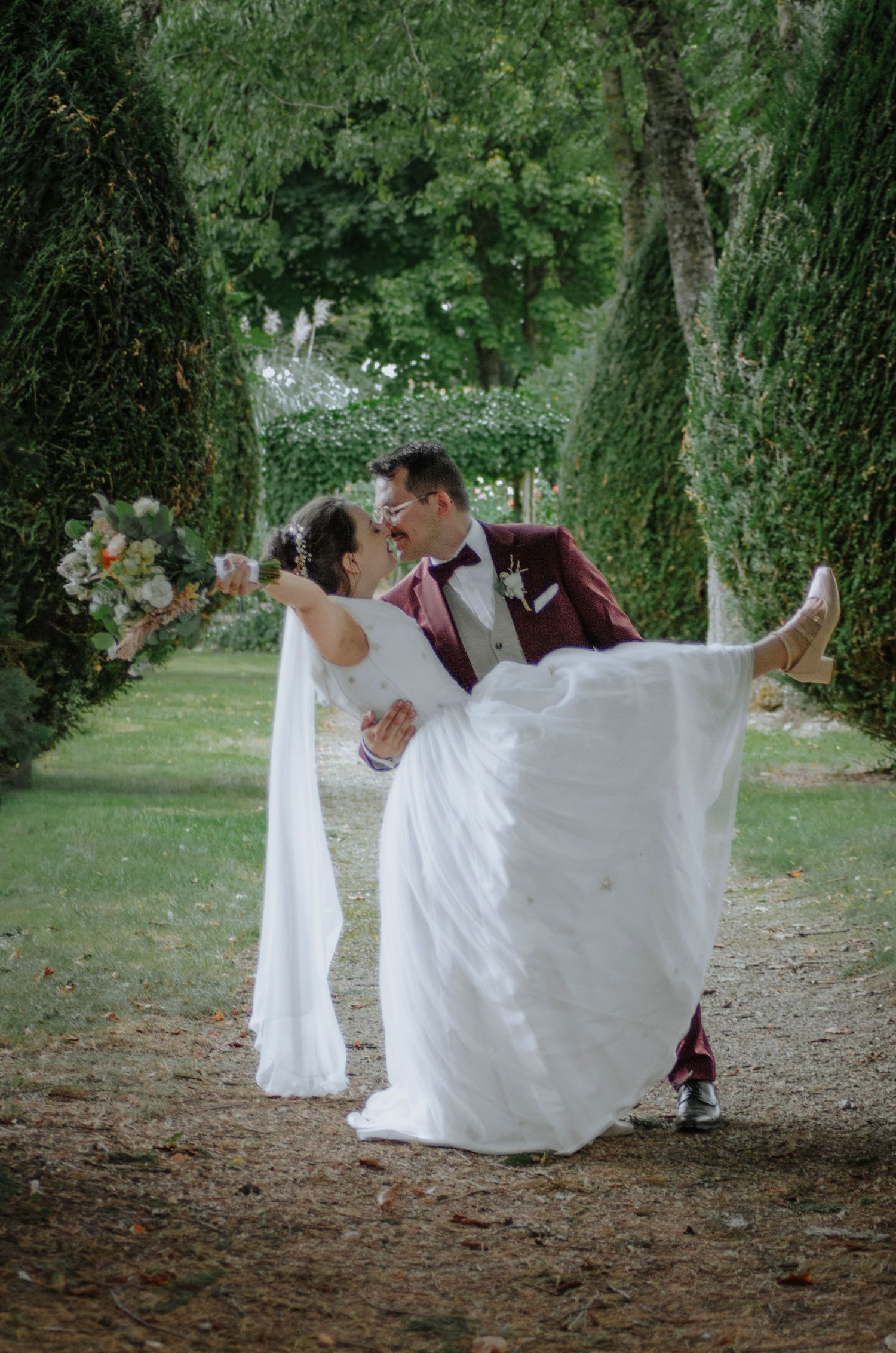 A couple in wedding attire walking through a scenic outdoor garden setting.