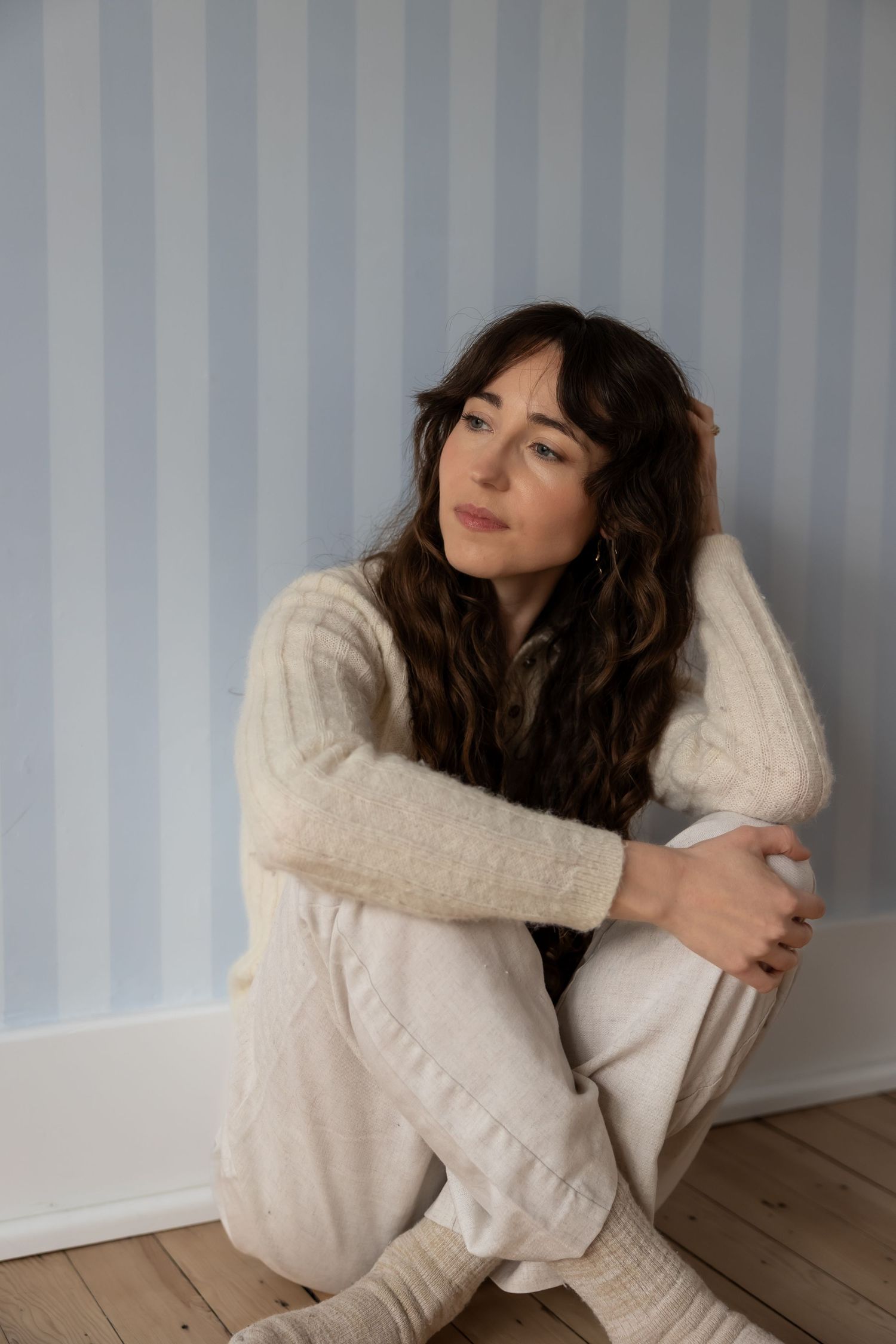 Person in cream knit sweater poses against gray wall in artistic portrait series.