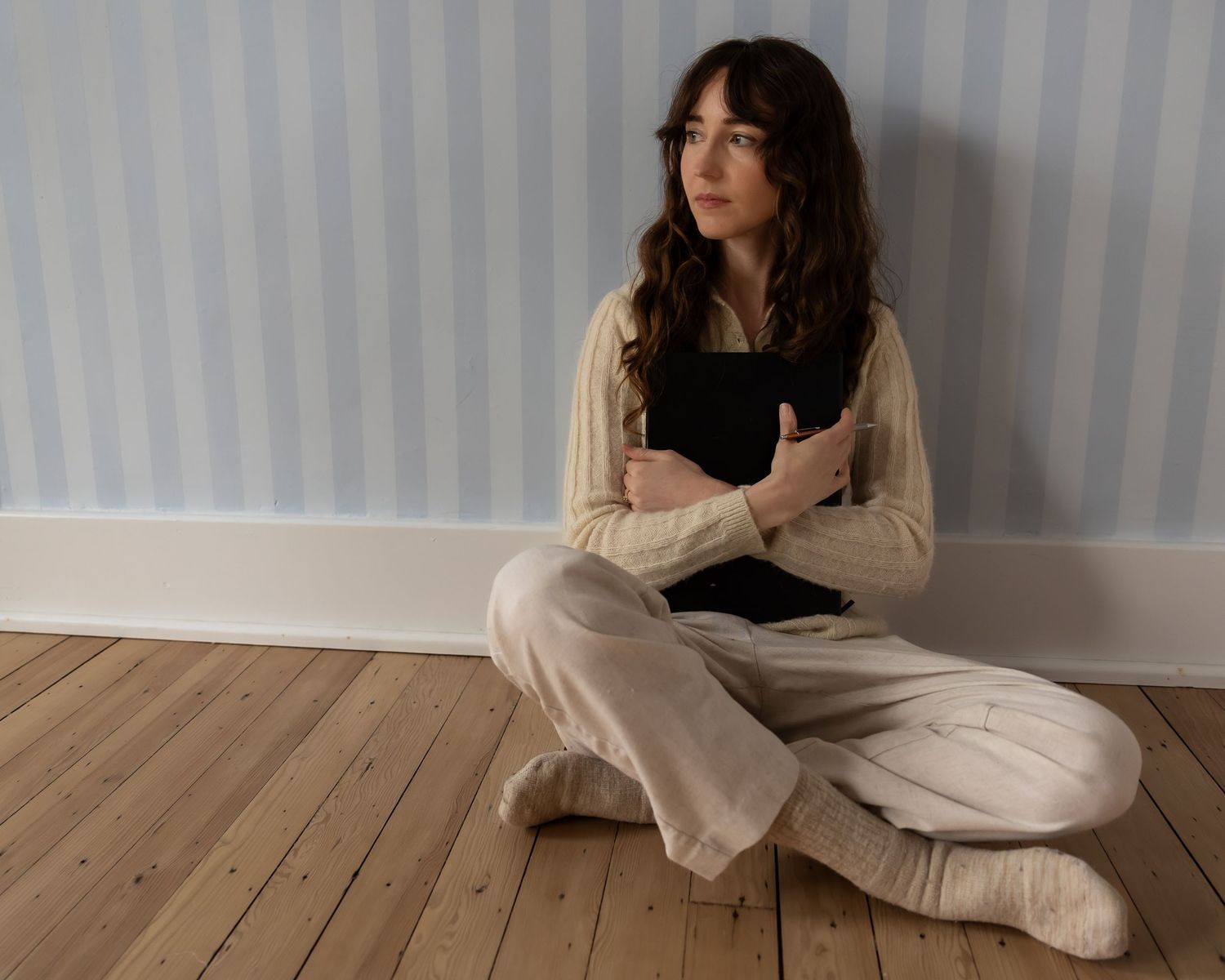 Woman in casual black and white clothing poses gracefully on wooden floor in minimalist setting.