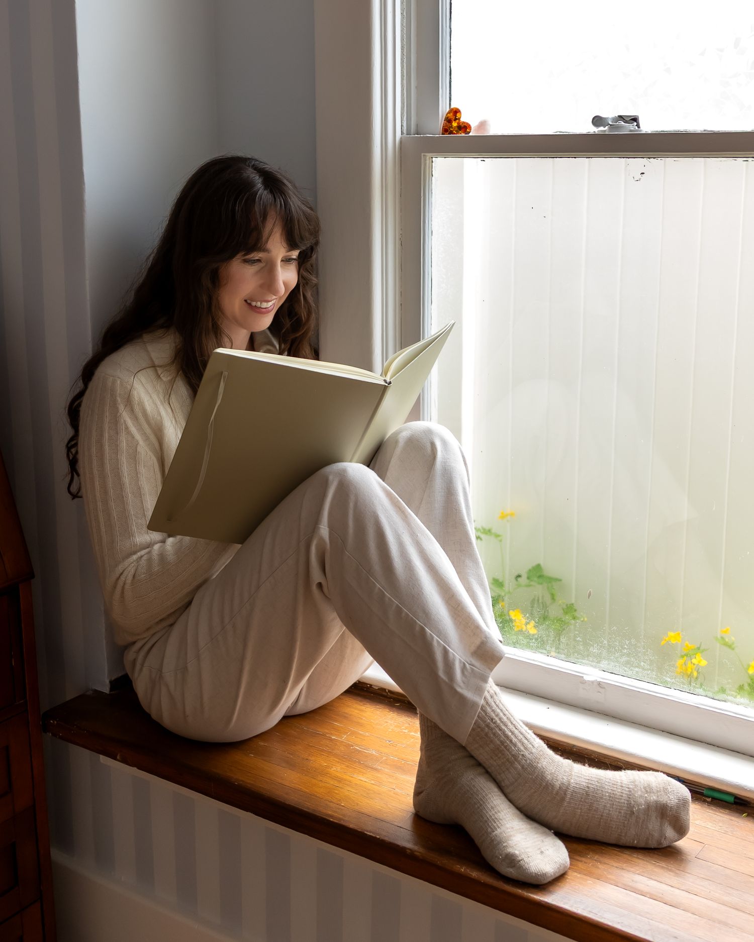 A person in a white sweater and socks sits comfortably on a window seat while reading a book in natural light.