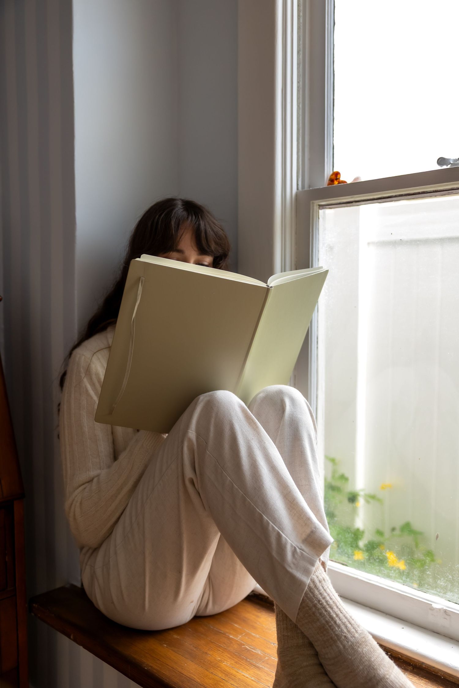 A person in white clothing sits curled up on a windowsill reading a book in soft natural light.