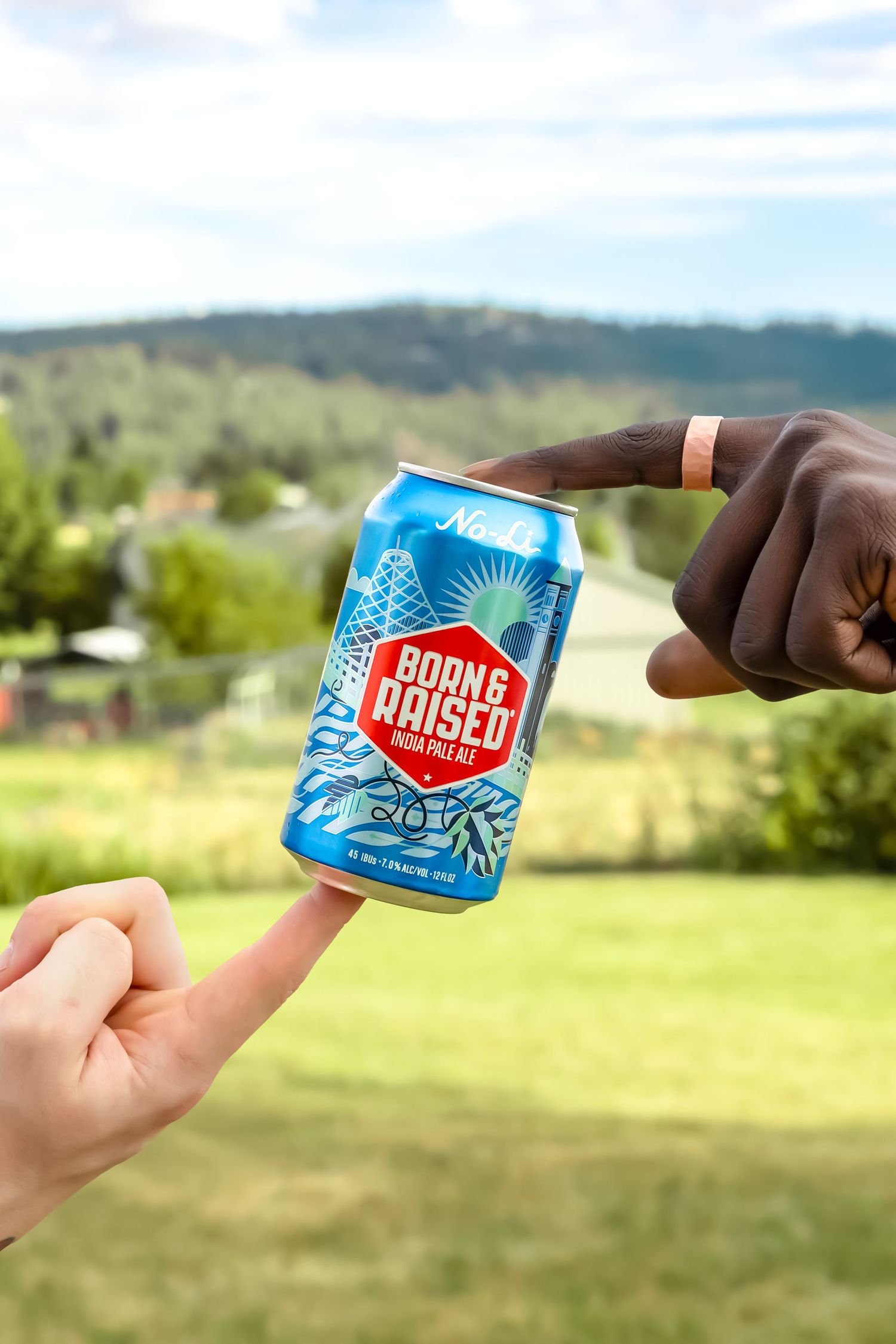 Two hands passing a blue can of Public Raised beer against a scenic mountain backdrop.