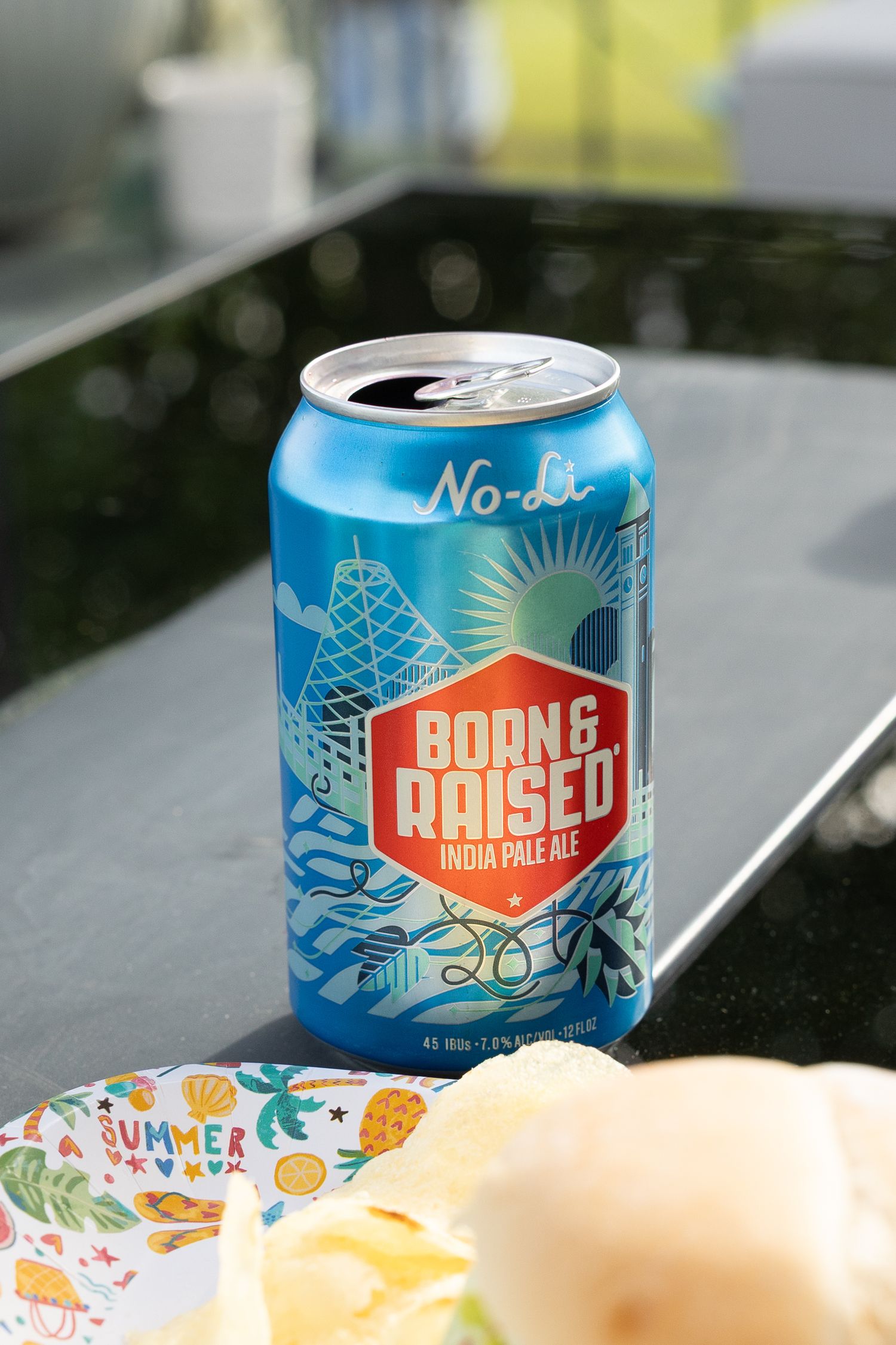 A blue can of Born & Raised India Pale Ale beer with white and orange design elements sits on a light surface.