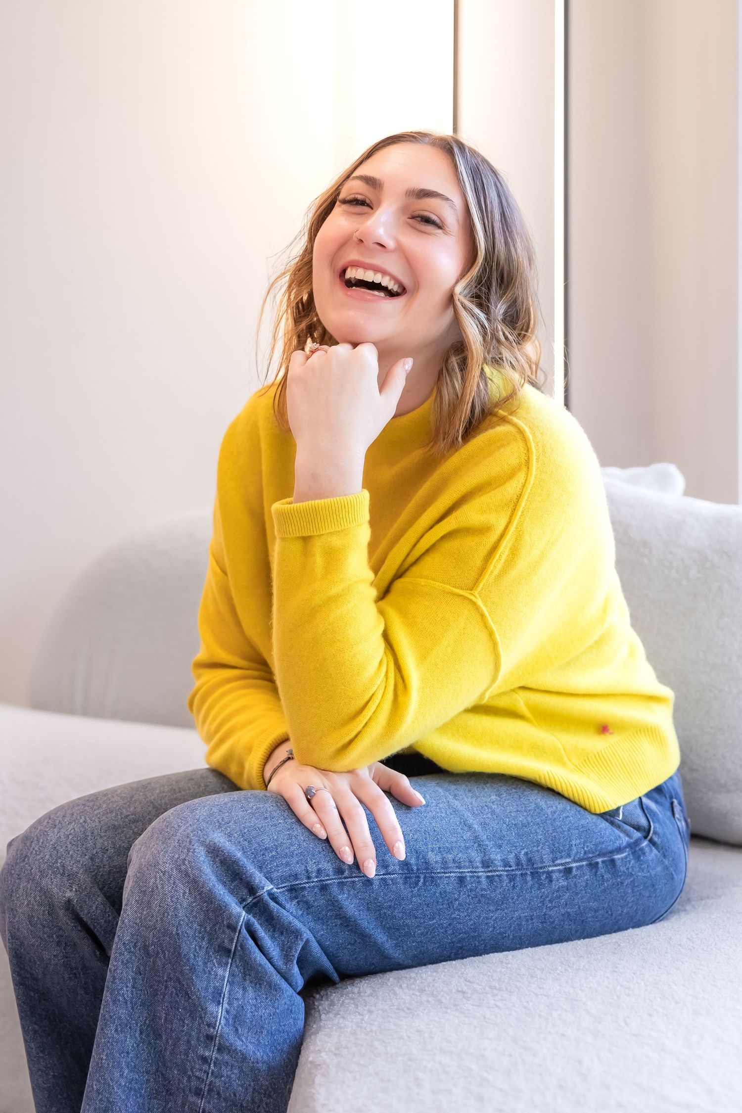 Person wearing bright yellow sweater and jeans lounging on gray couch while laughing joyfully.