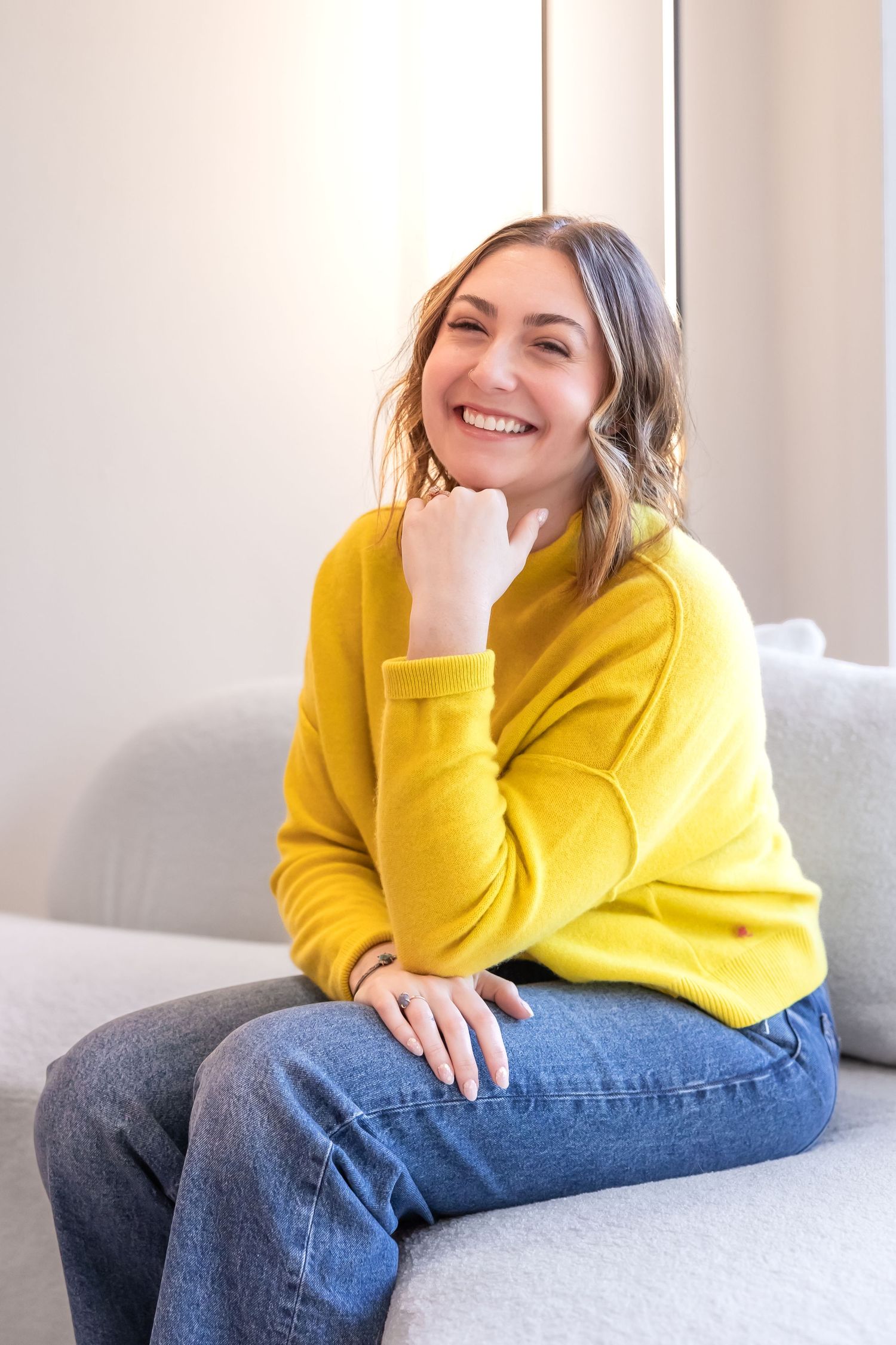 Woman in bright yellow sweater and jeans lounges casually on gray couch showing different relaxed poses.