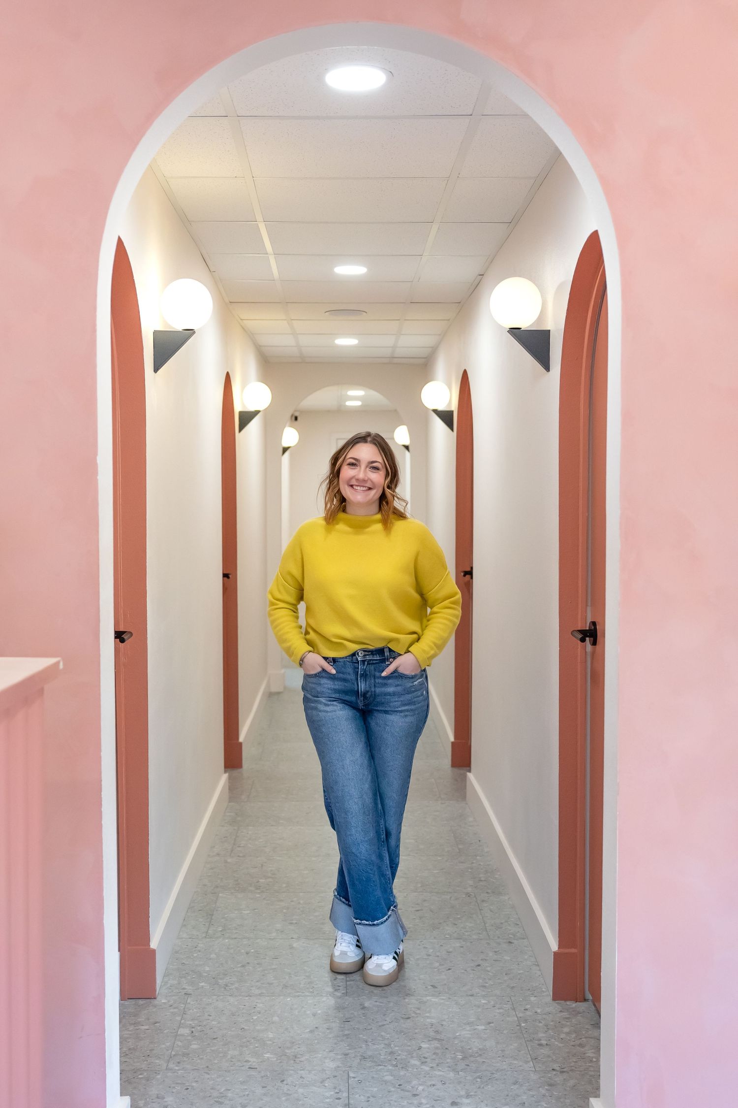 Person in yellow sweater and jeans stands in white hallway with pink walls and arched doorways.