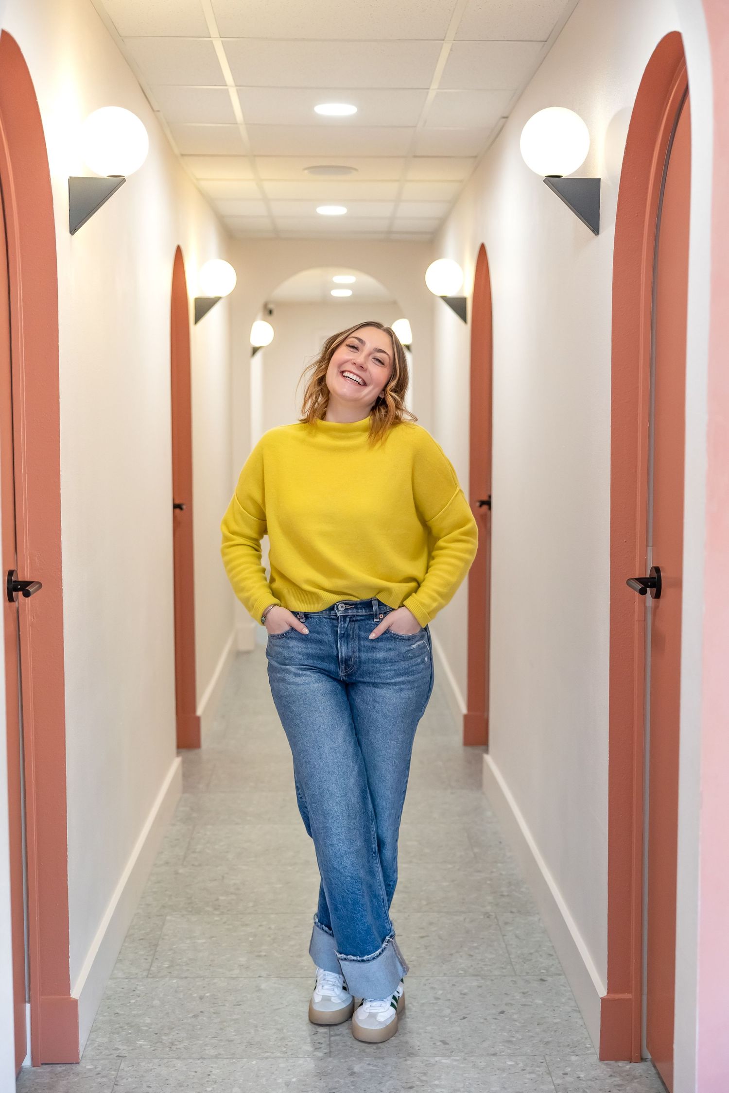 Person in bright yellow sweater and jeans poses playfully in a long hallway with pink-toned walls and white ceiling.