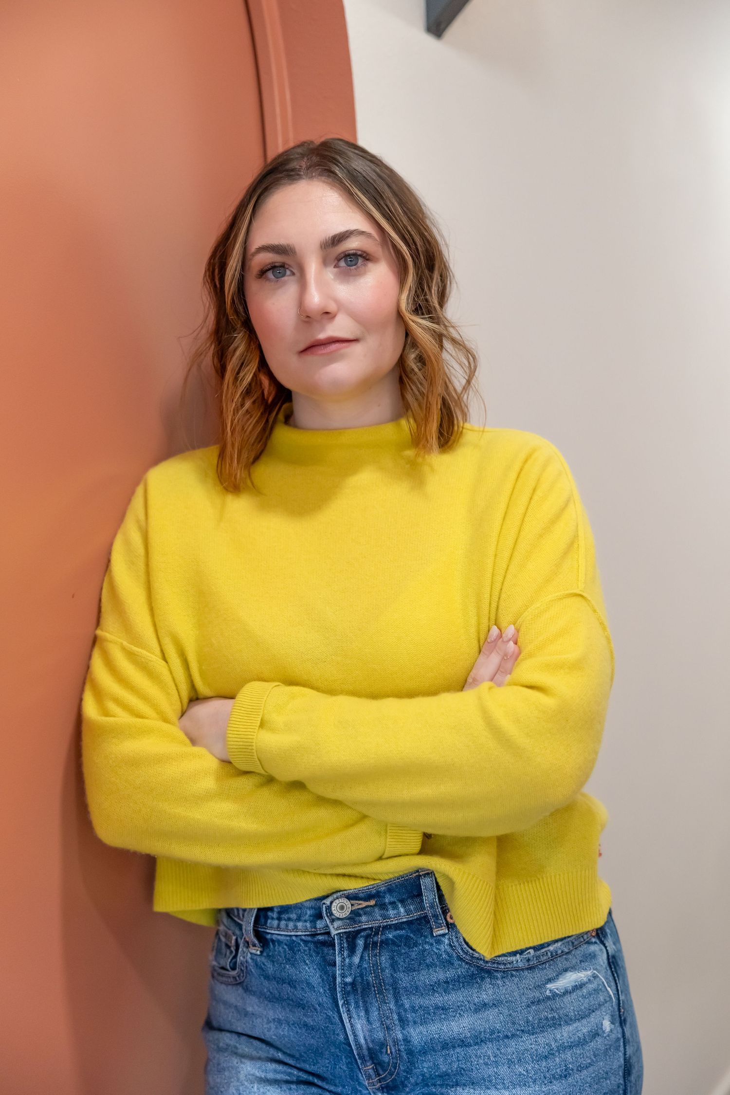 Person wearing bright yellow sweater and blue jeans stands with arms crossed against coral and white wall.