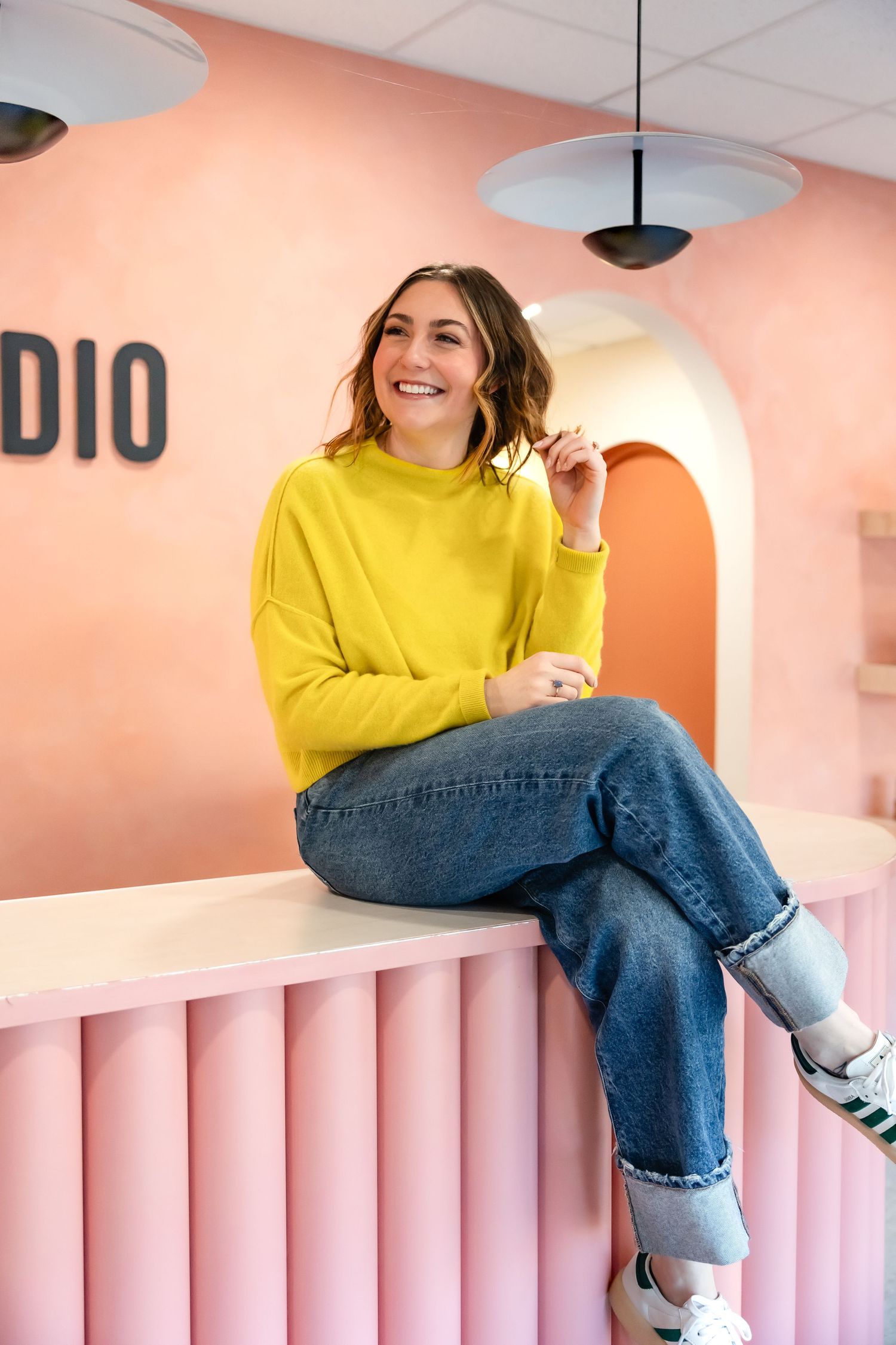Person wearing yellow sweater and jeans poses playfully on pink striped wall installation.