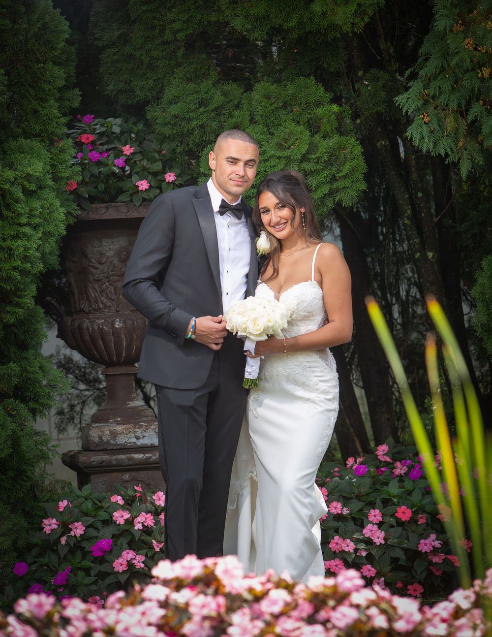 A couple in formal wedding attire poses in a garden setting with pink flowers and a decorative stone urn.
