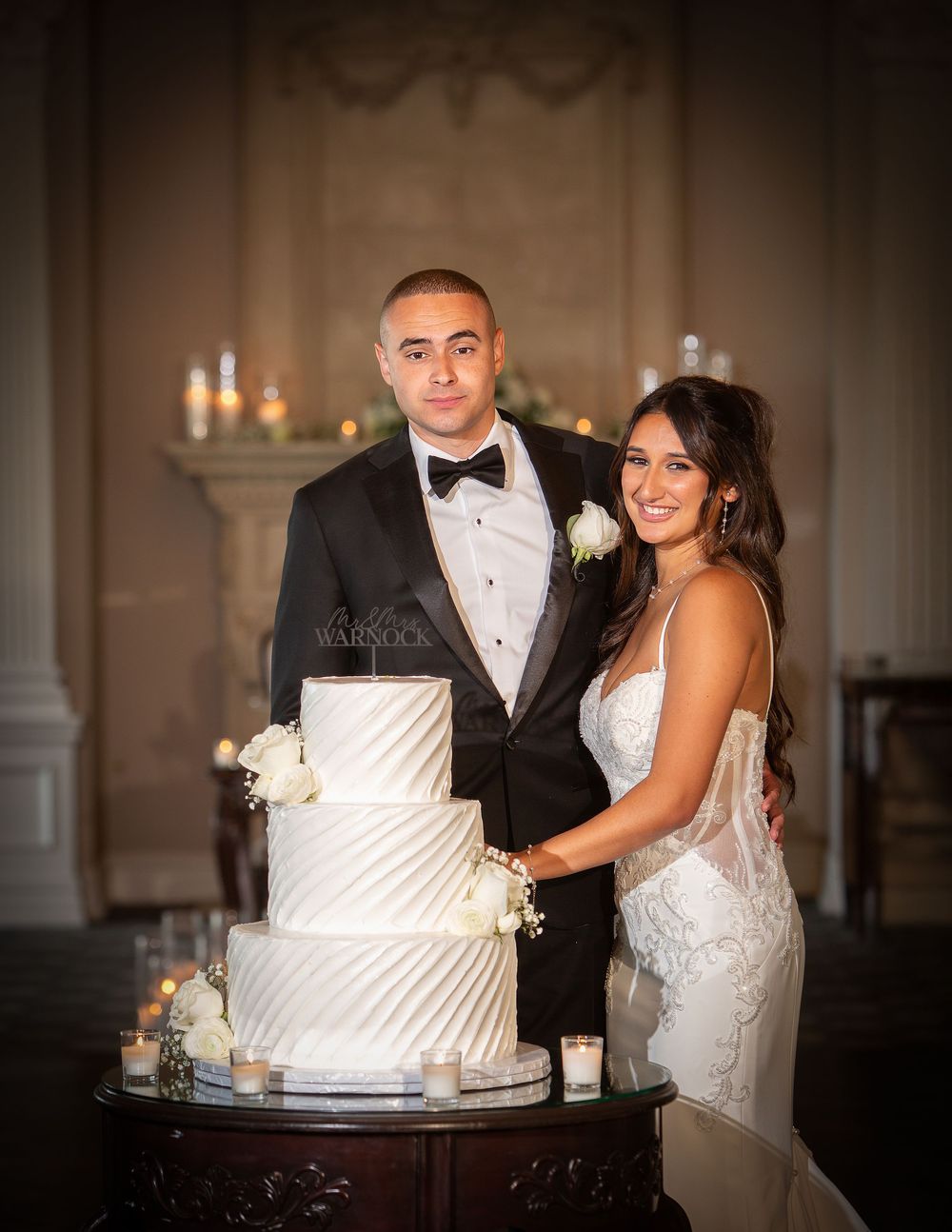 A bride and groom pose together with their elegant white tiered wedding cake decorated with diagonal stripes.