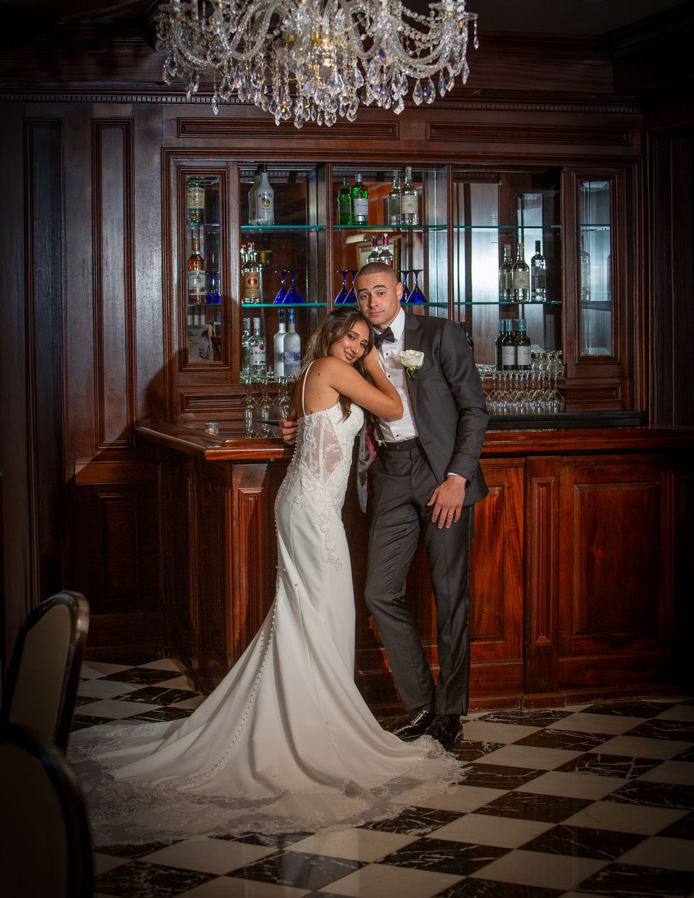 A romantic couple poses in an elegant wood-paneled ballroom with a crystal chandelier and checkered floor.