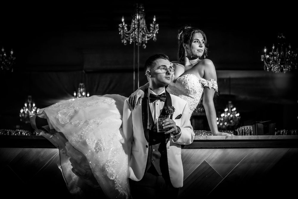 A black and white wedding photo shows a couple posing dramatically at a bar with elegant chandeliers in the background.
