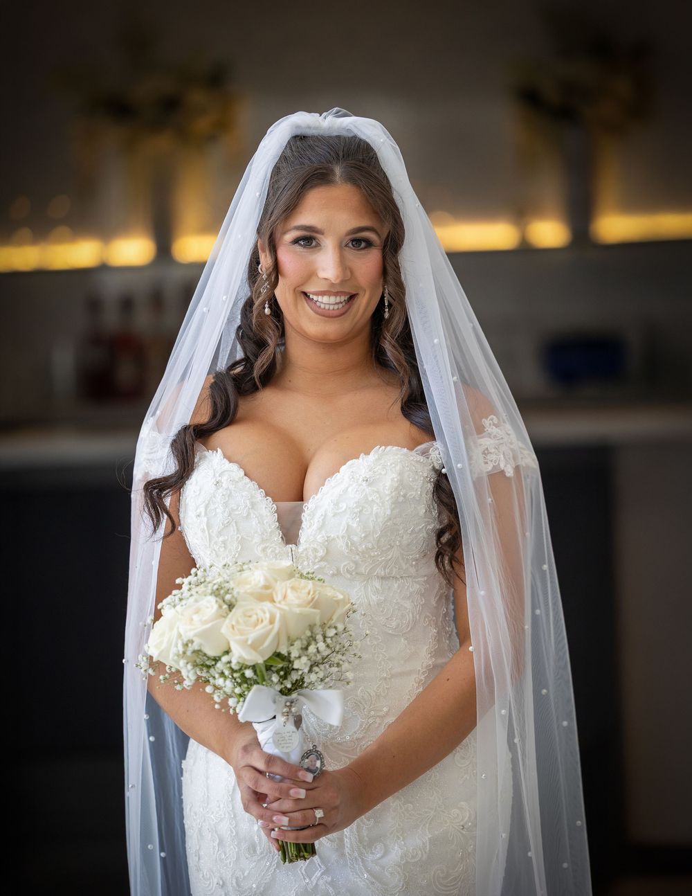 A bride in a white lace wedding dress with a long veil holds a white rose bouquet against a blurred background.
