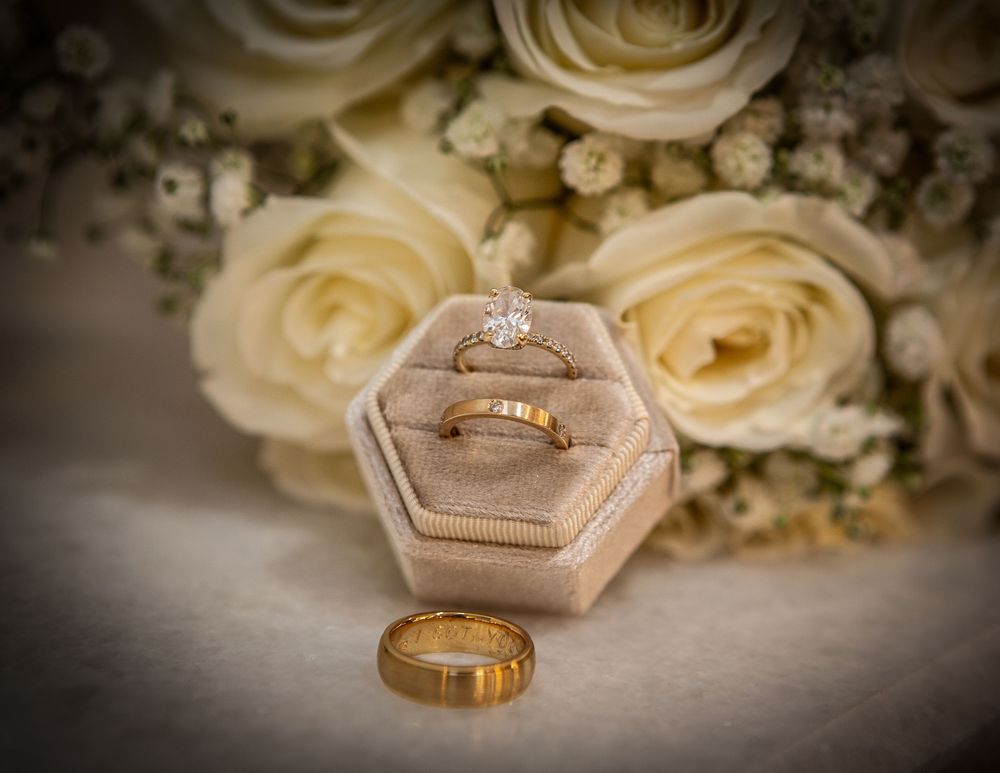 Wedding rings displayed on a hexagonal ring box against a backdrop of soft cream-colored roses and white floral arrangements.
