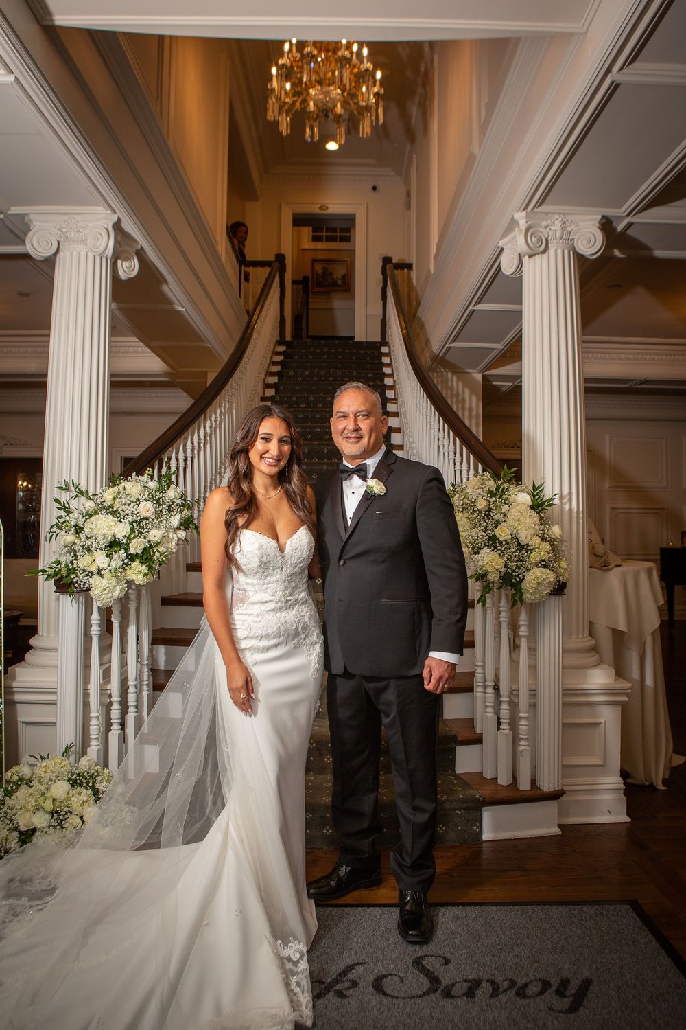 Elegant wedding couple poses in grand ballroom with white columns and floral arrangements at The Savoy.