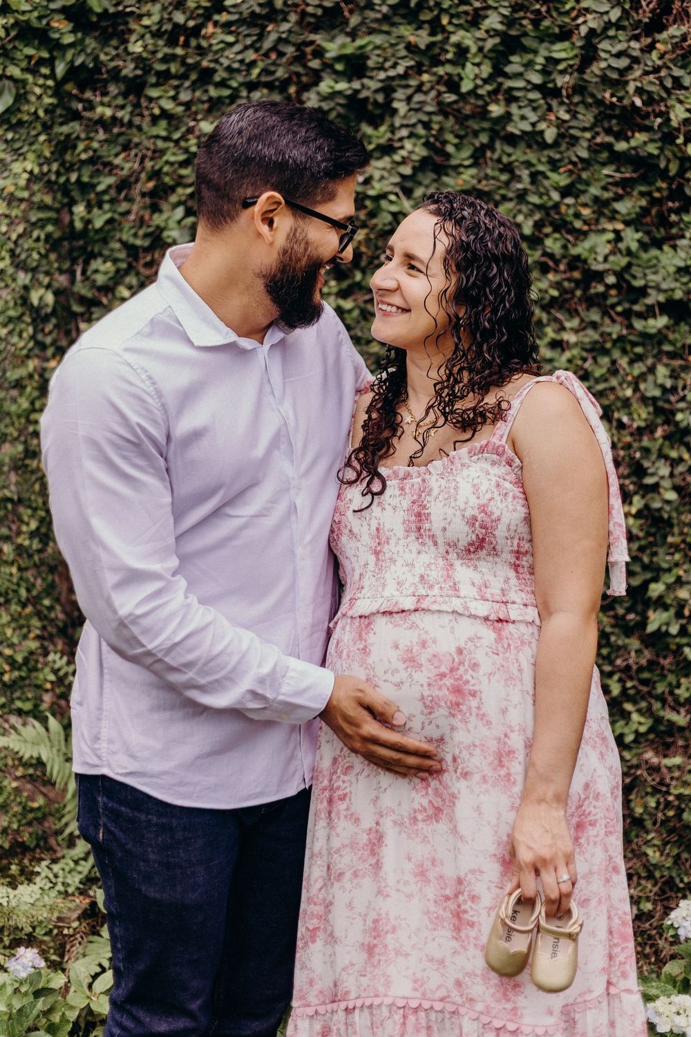 Couple shares intimate moment in garden setting while holding tiny shoes.