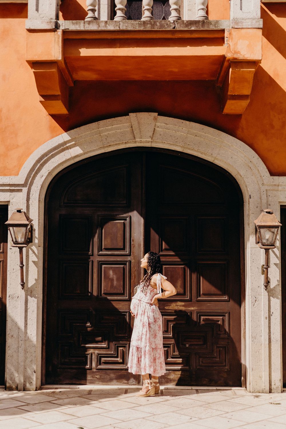 Woman in floral dress poses under historic stone archways in a Mediterranean style building.