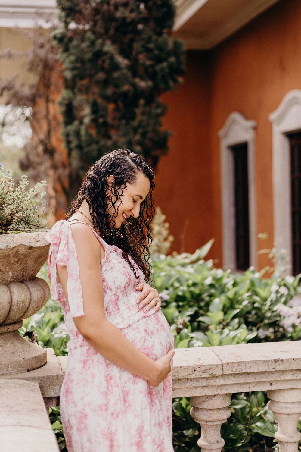 A maternity session featuring a pink floral dress against a terracotta colored wall.