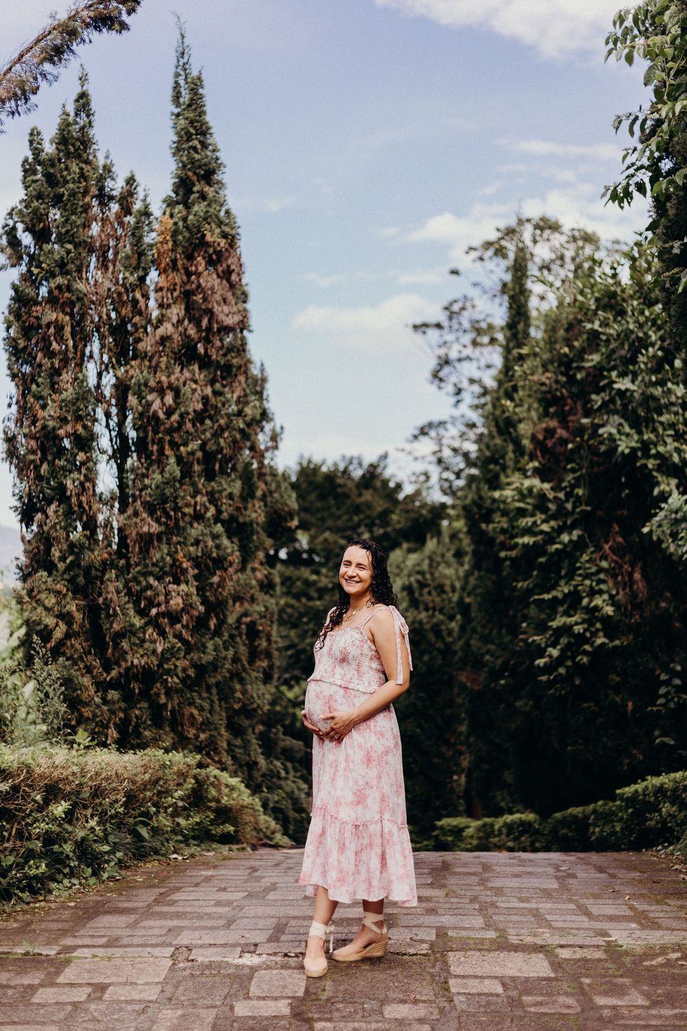 A woman in a floral pink dress stands on stone pavement surrounded by tall cypress trees in a garden setting.