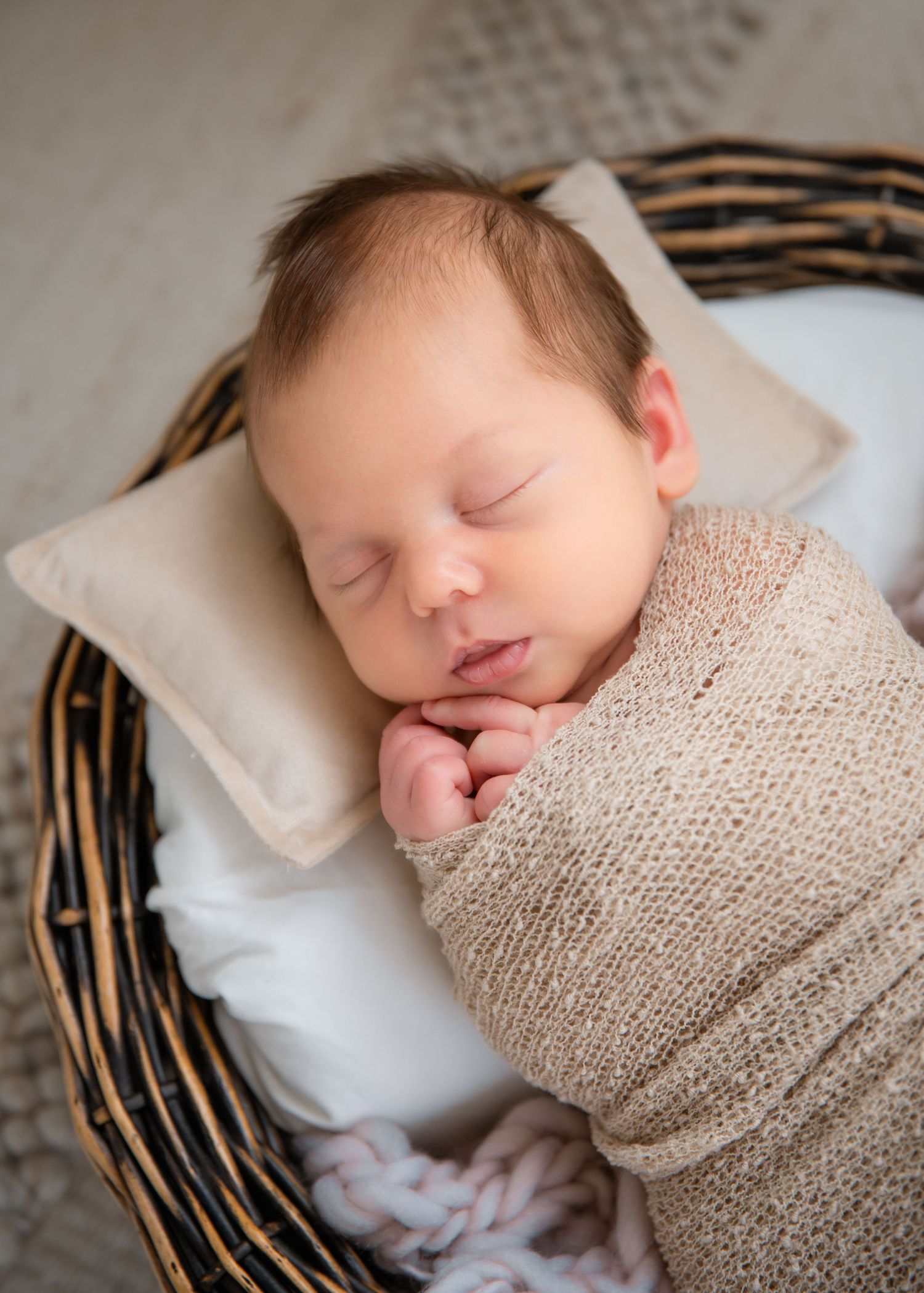 A sleeping newborn baby wrapped in beige knit blanket lying on white pillow in woven basket.
