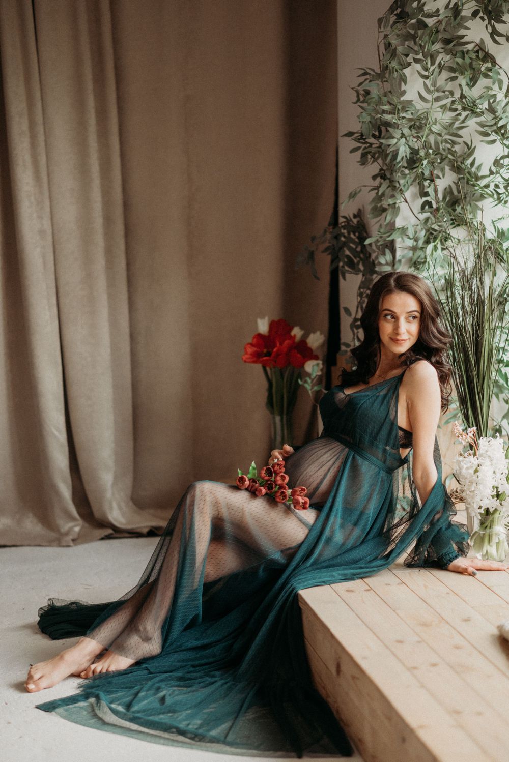 Woman in teal flowing dress posing elegantly near indoor plants and curtains.