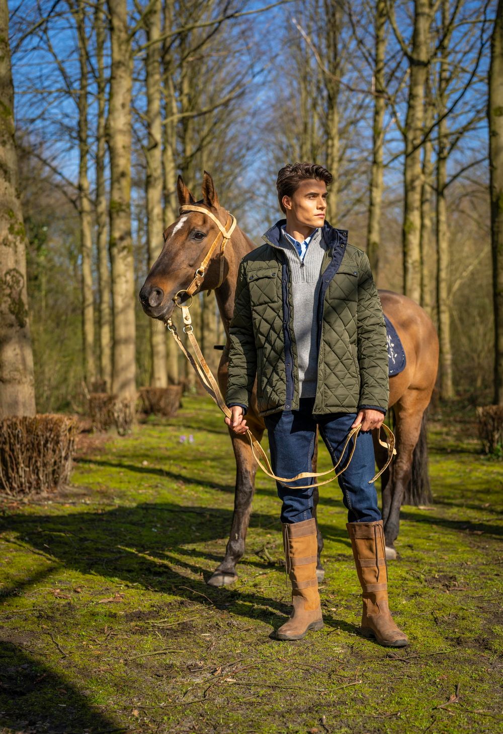 A beautiful brown horse stands with its handler in a wooded area on a sunny spring day.