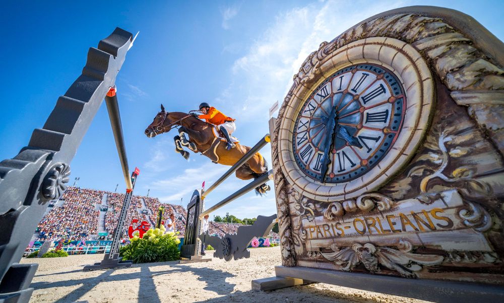 Horse and rider leaping over obstacle near large decorative clock structure.