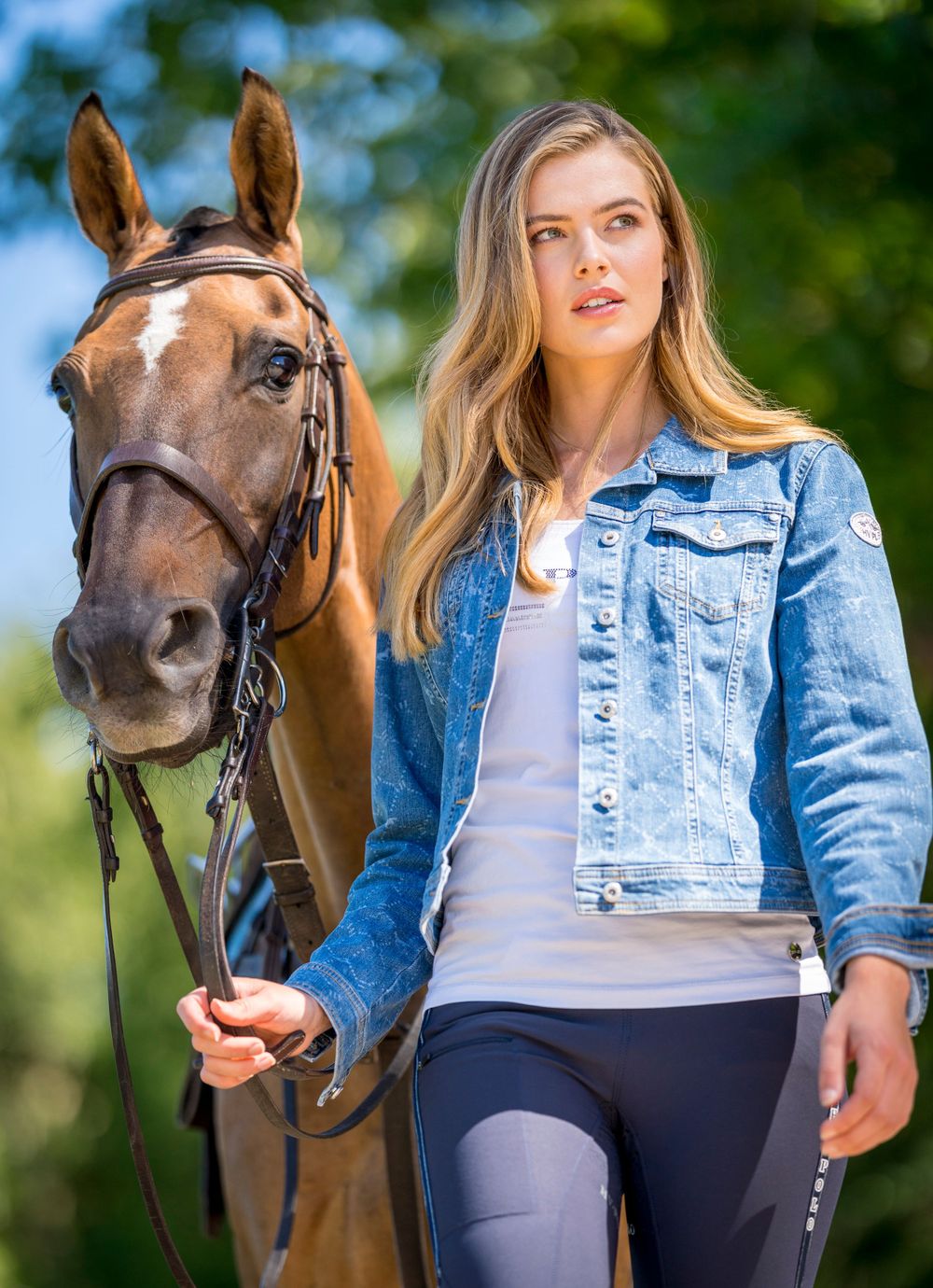 Equestrian with long blonde hair wearing denim jacket poses with brown horse outdoors.