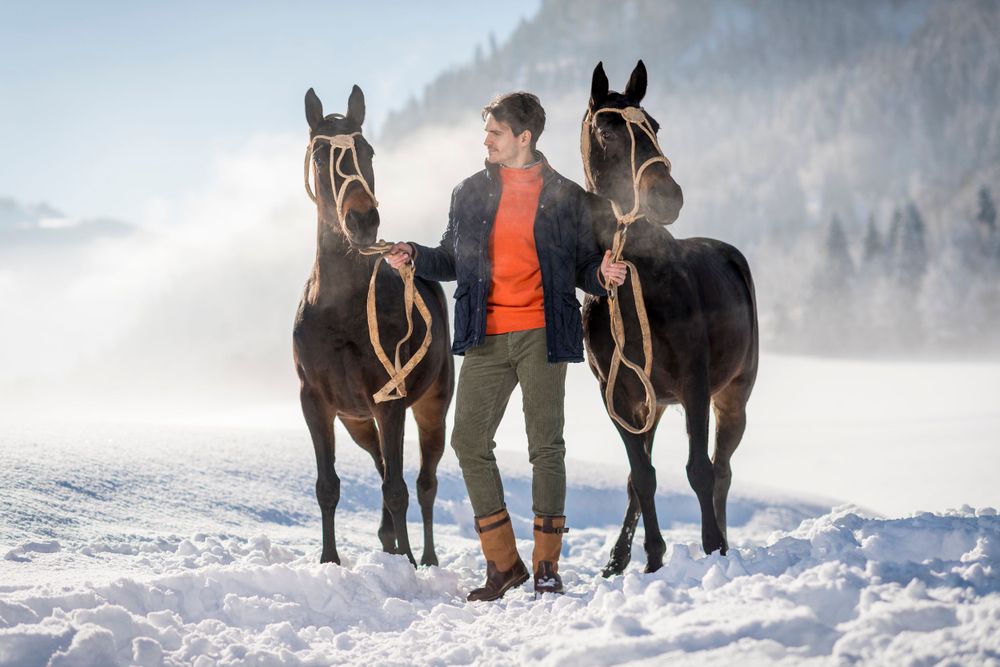 Two dark horses stand beside a person wearing orange and gray in a snowy winter landscape with misty mountains.