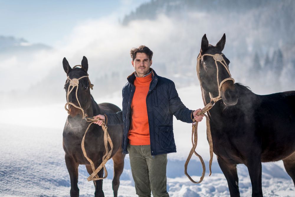 Two elegant horses flank a figure in an orange sweater and navy jacket against a snowy mountain backdrop.