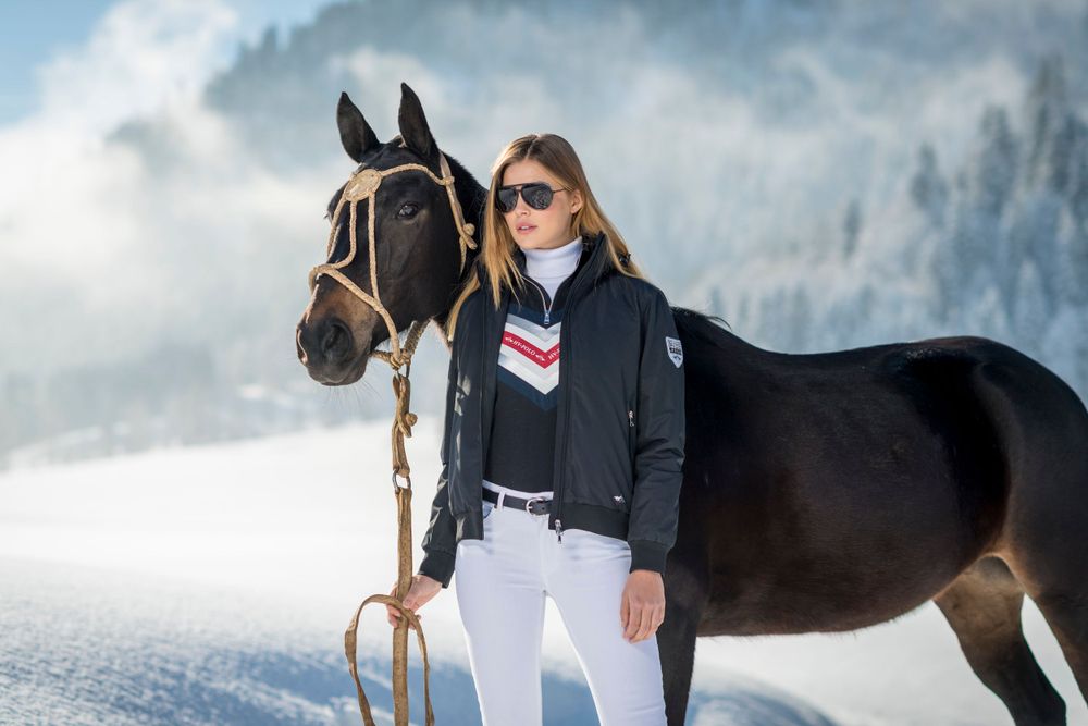 Person in winter attire stands with black horse against snowy mountain landscape.